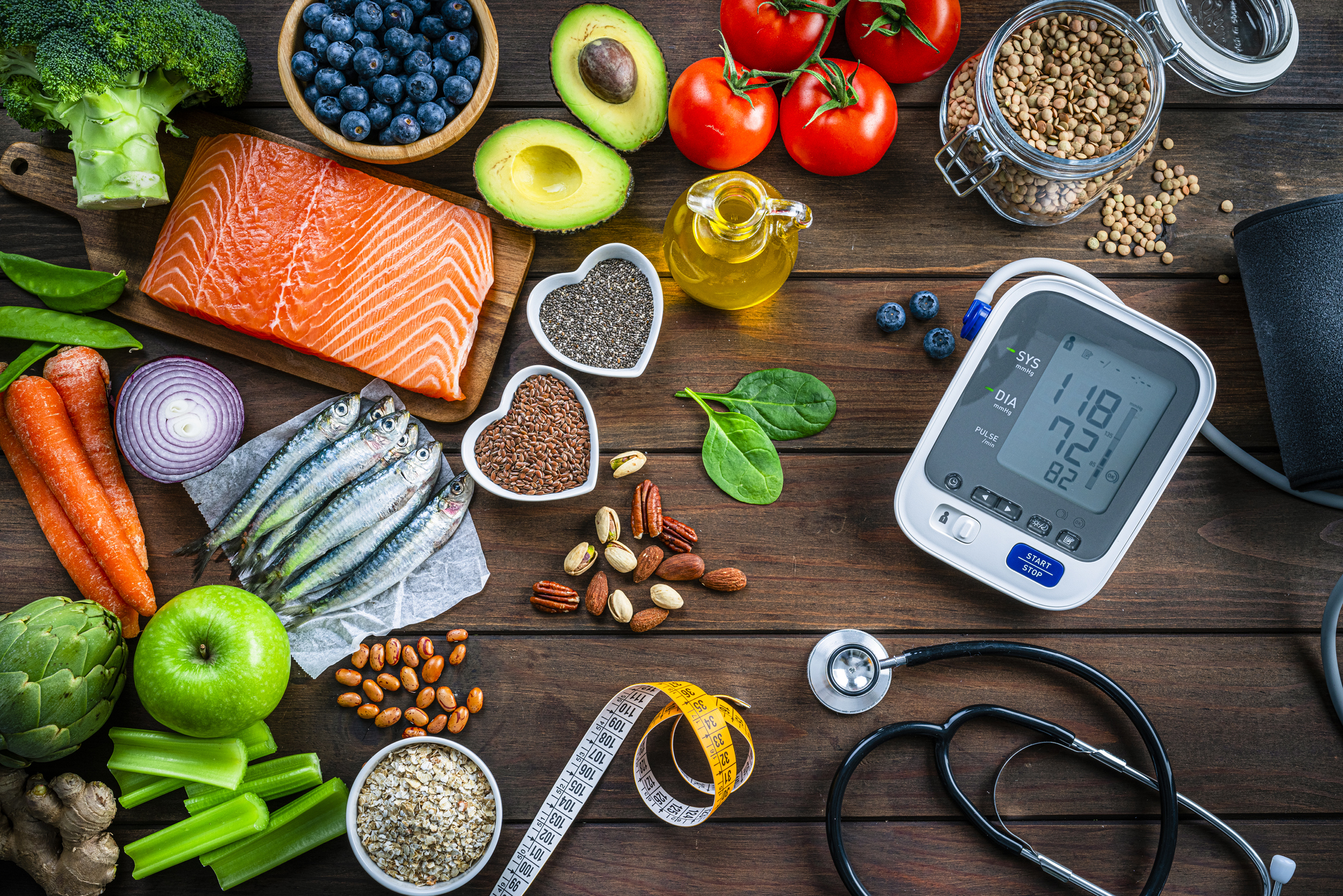 Healthy food for heart care: overhead view of healthy food rich in Omega-3 and antioxidants and a blood pressure monitor, tape measure and stethoscope shot on wooden table. The composition includes a salmon fillet, sardines, avocado, extra virgin olive oil, brown lentils, celery, artichoke blueberries, celery, carrots, brown lentils, asparagus, artichoke, broccoli, flax seeds, chia seeds and some nuts like almonds, pistachio and pecan. High resolution 42Mp studio digital capture taken with SONY A7rII and Zeiss Batis 40mm F2.0 CF lens
