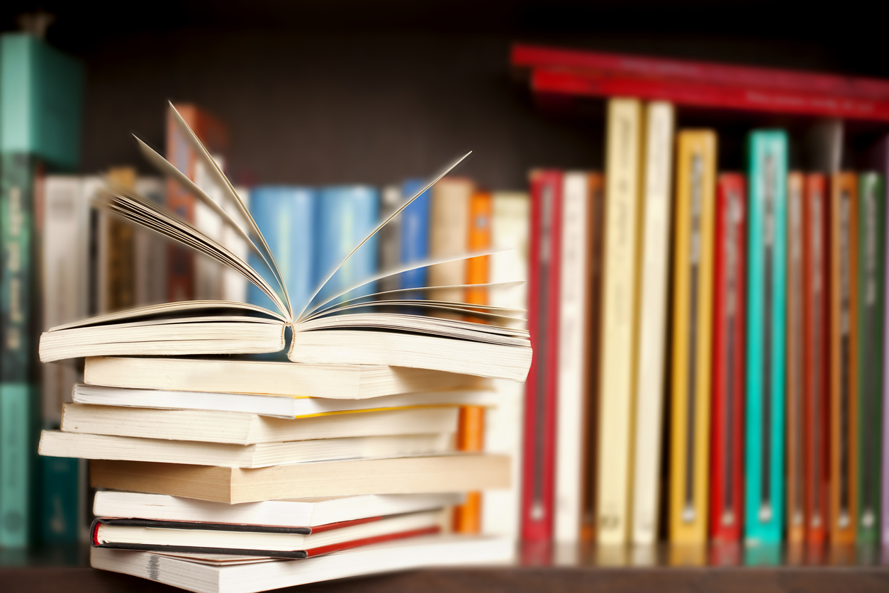 Stack of books on a wooden library shelf, one of them open on top, multicolored book spines background.