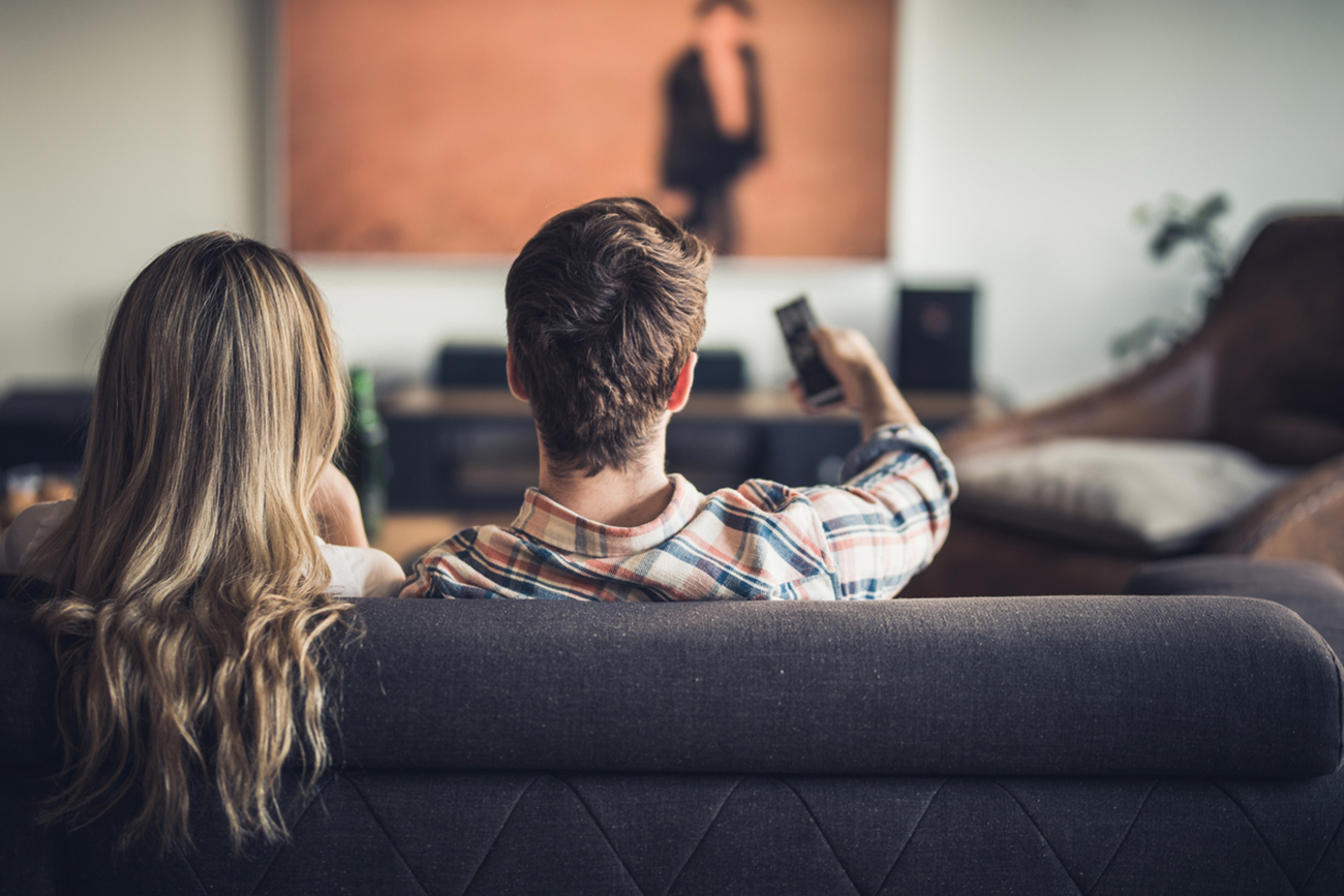 Back view of a couple watching TV on sofa at home. Man is changing channels.