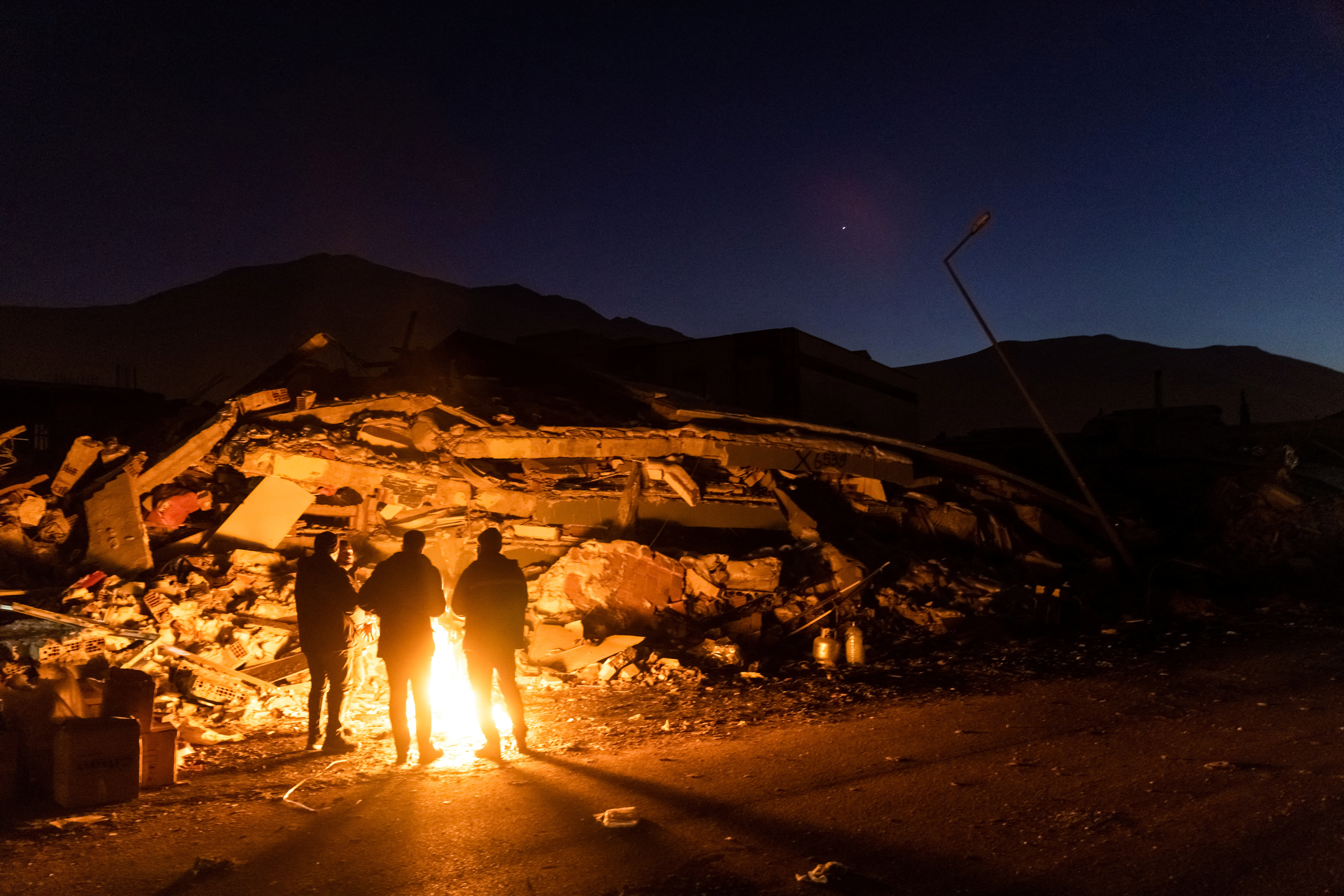 People stand by the fire next to remains of a destroyed building in the aftermath of a deadly earthquake in Elbistan, Turkey February 14, 2023. REUTERS