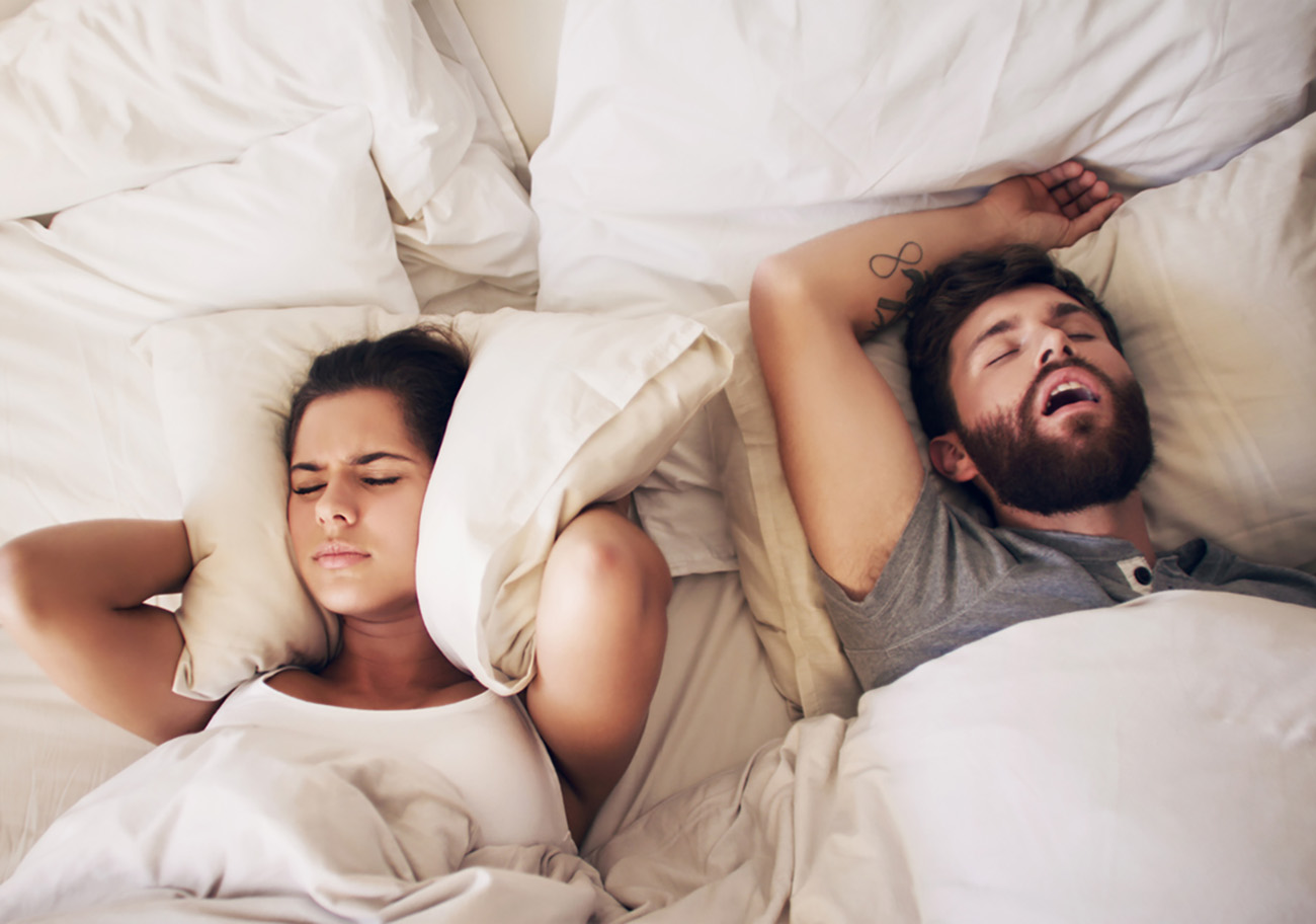 Shot of a young woman covering her ears with a pillow while her husband snores in bed