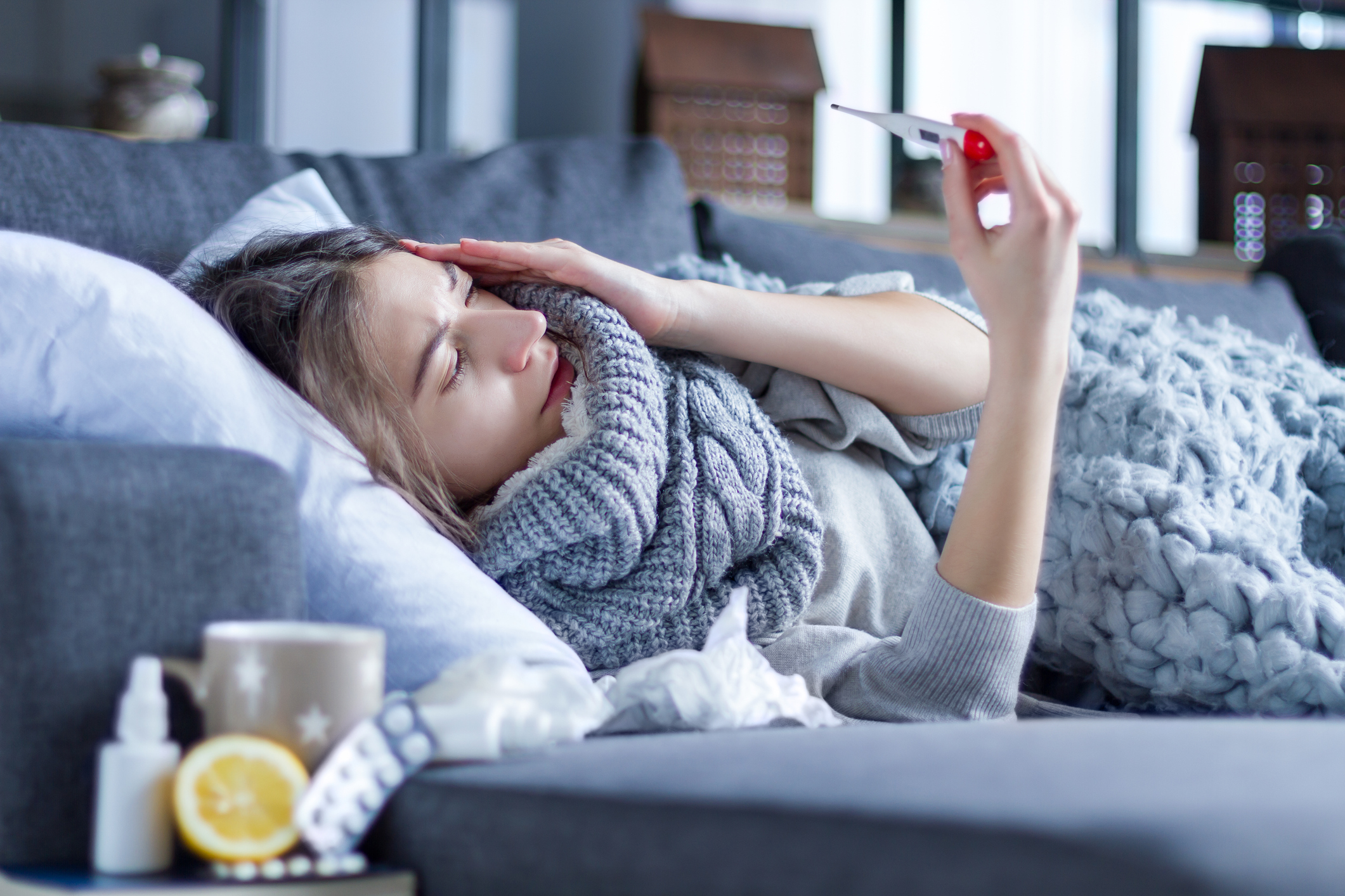 Sick exhausted girl in scarf is lying in bed wrapped in blanket. Young woman with fever and headache is measuring temperature with thermometer, treated at home. Winter cold and flu concept.