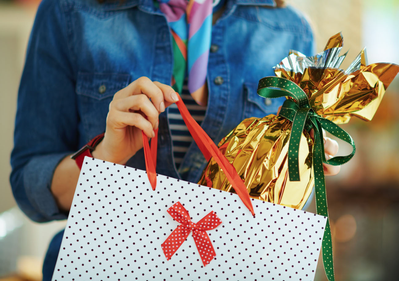 Closeup on woman in the house in sunny spring day taking out wrapped in gold foil big easter egg from white polka dot shopping bag.