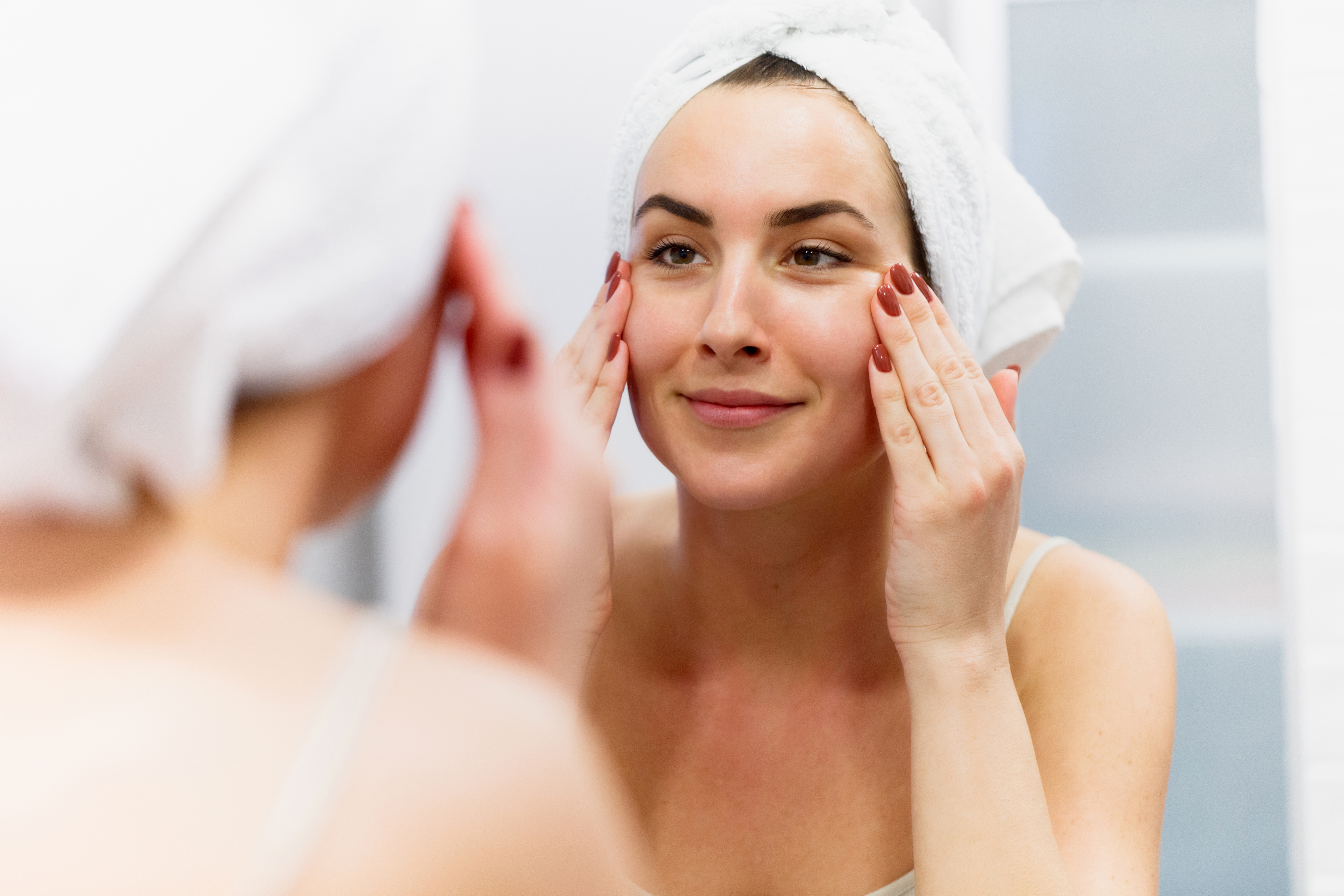 Smiling woman with towel on wet hair reflecting in the bathroom mirror and tightening her cheeks, pulling skin back