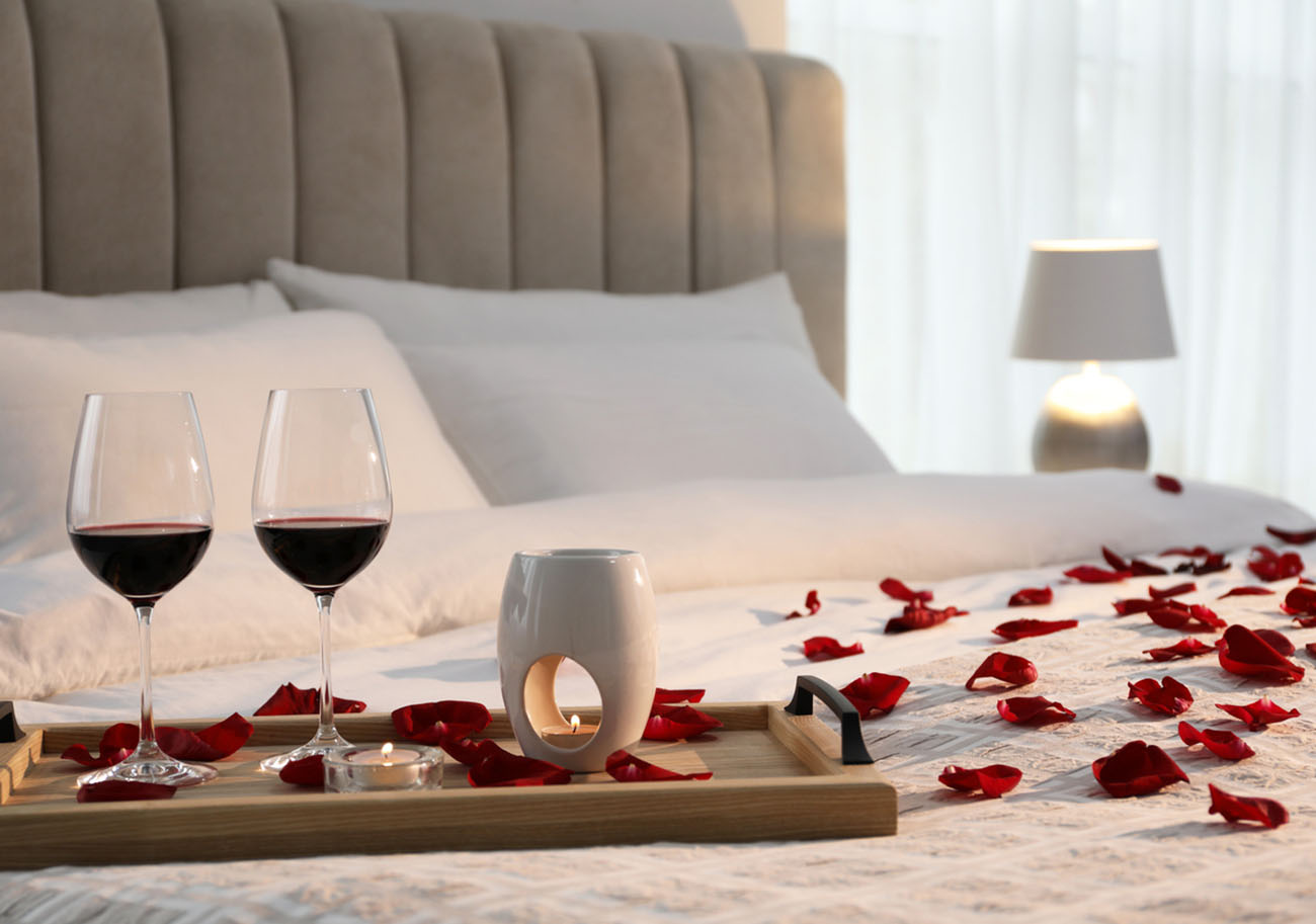 Tray with glasses of red wine, candles and rose petals on bed in room