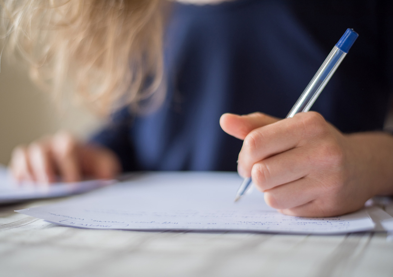 The photo shows a close up of a woman who is writing a text with her hand and a pen on paper. This handwriting background image is taken at a table.