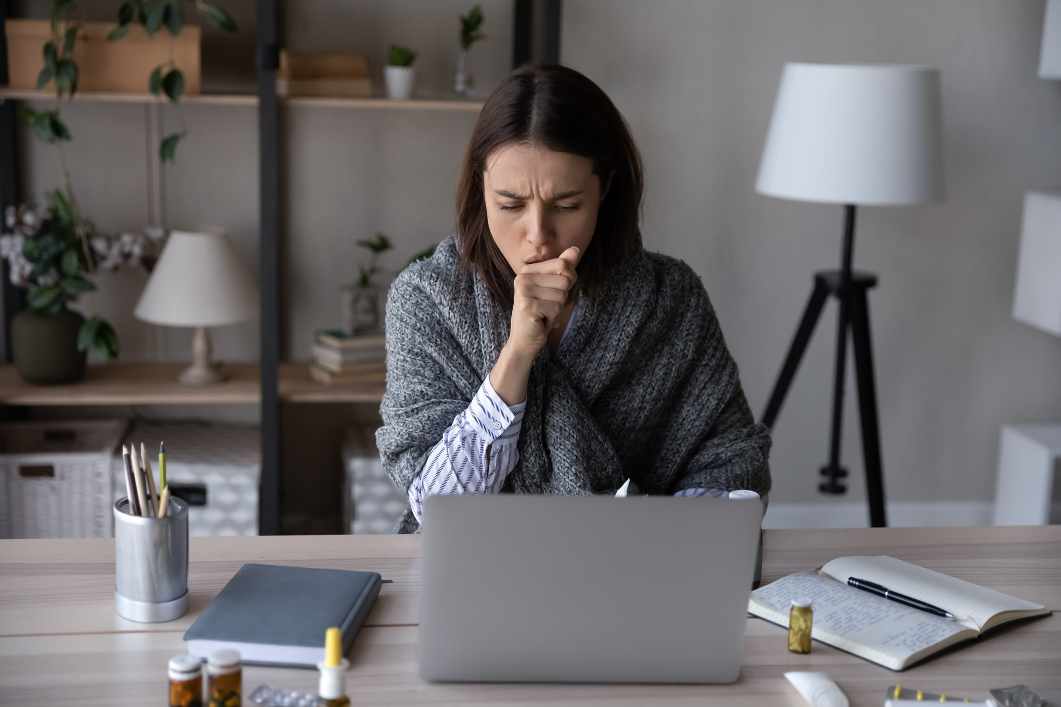Unhealthy young Caucasian woman sit at desk at home office work online on computer suffer from covid-19 virus. Sick female busy at laptop cough struggle with coronavirus or flu at workplace.