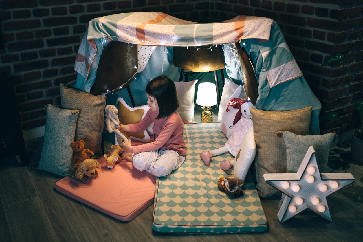 Adorable girl playing alone with teddies in a tent made of bed sheets and chairs