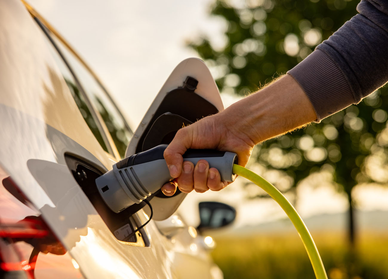 Hand of man inserting a power cord into an electric car for charging ecofriendly vehicle on green landscape