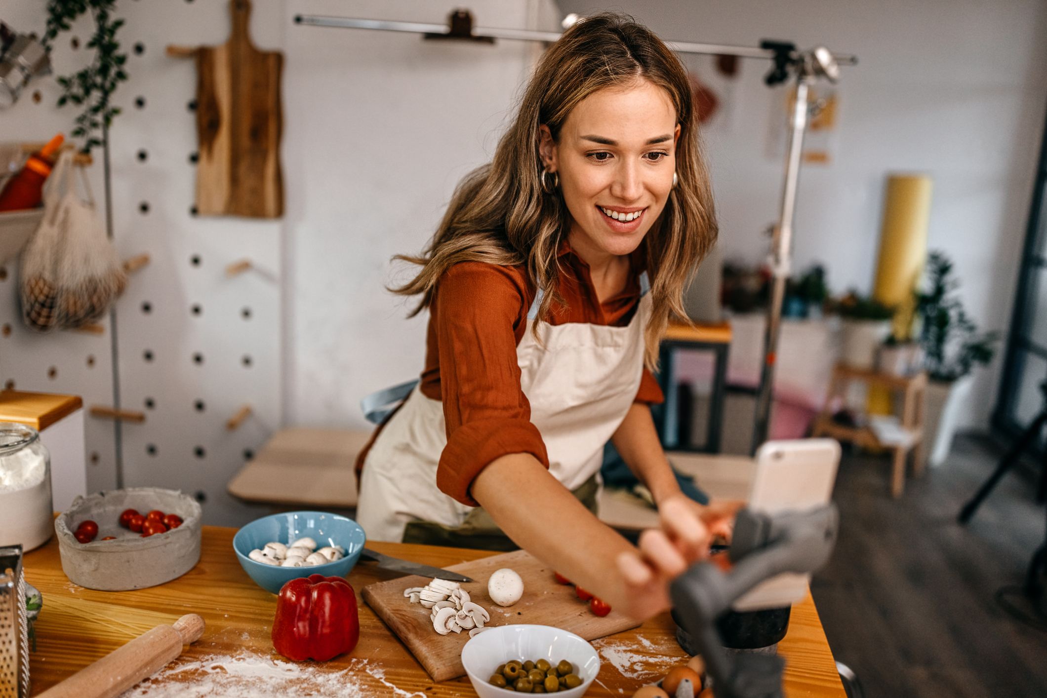Smiling and cheerful young female food blogger wearing apron speaking to audience during live streaming on social media while preparing pizza and chopping vegetables in modern kitchen studio