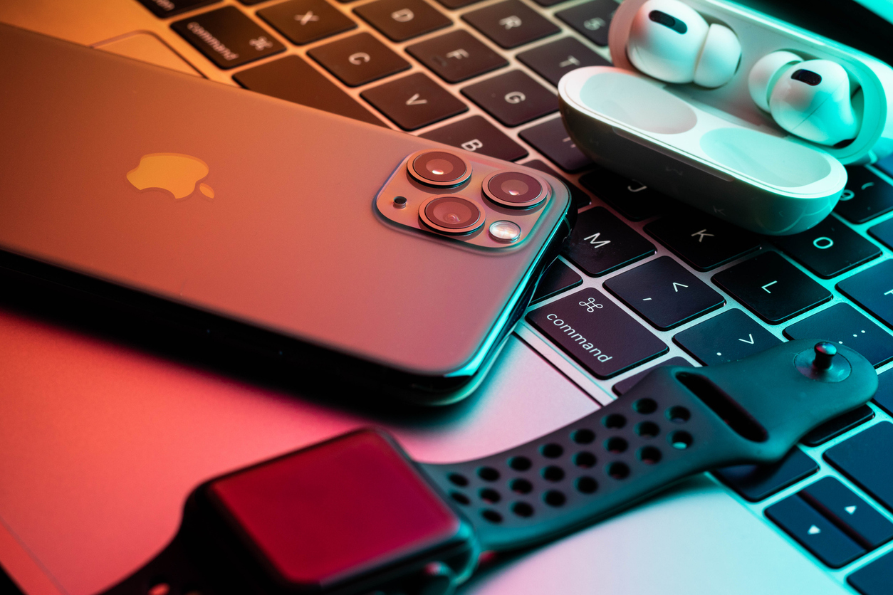 iPhone 11 Pro with AirPods Pro and Apple Watch with a Macbook in the background with teal and orange glow.