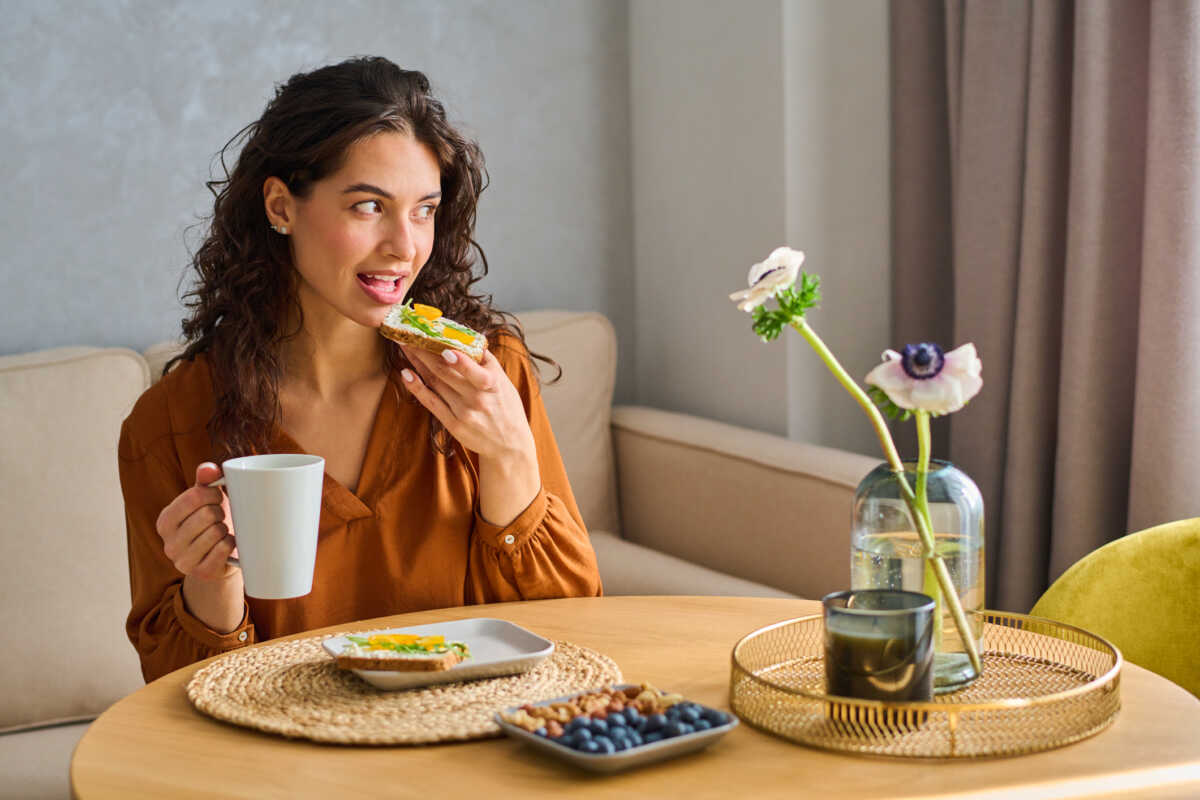 Young female looking through window while having breakfast by kitchen table served with sandwiches, blueberries and nuts on plates