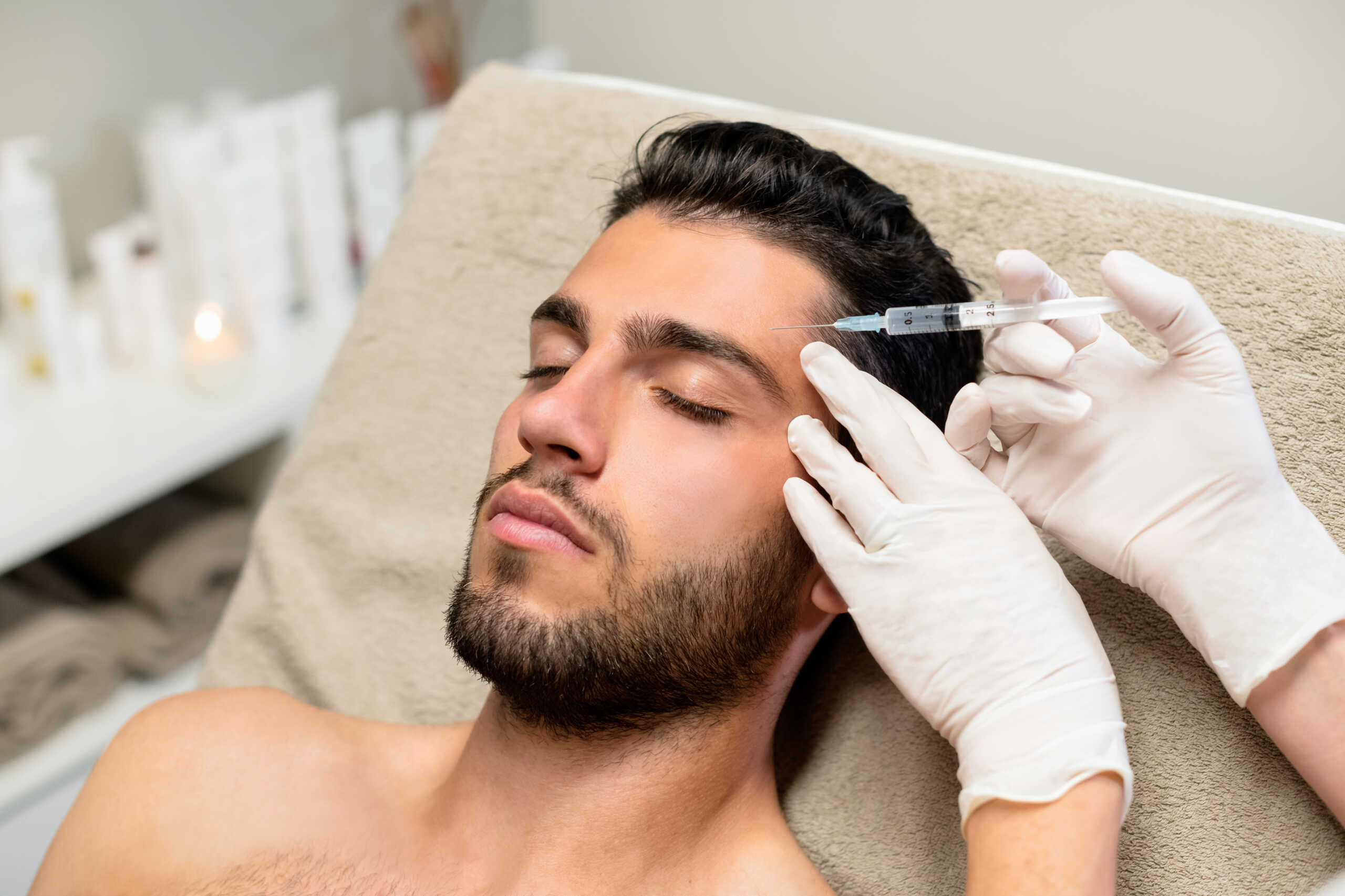 From above anonymous beautician in latex gloves injecting botulinum toxin into forehead of bearded young male client during cosmetology session in beauty studio