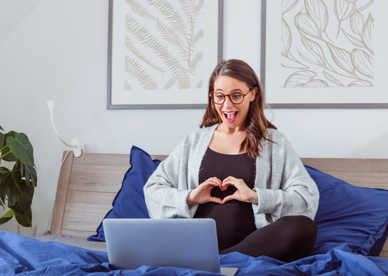 Excited pregnant woman sitting cross legged on the bed having video call, using laptop.