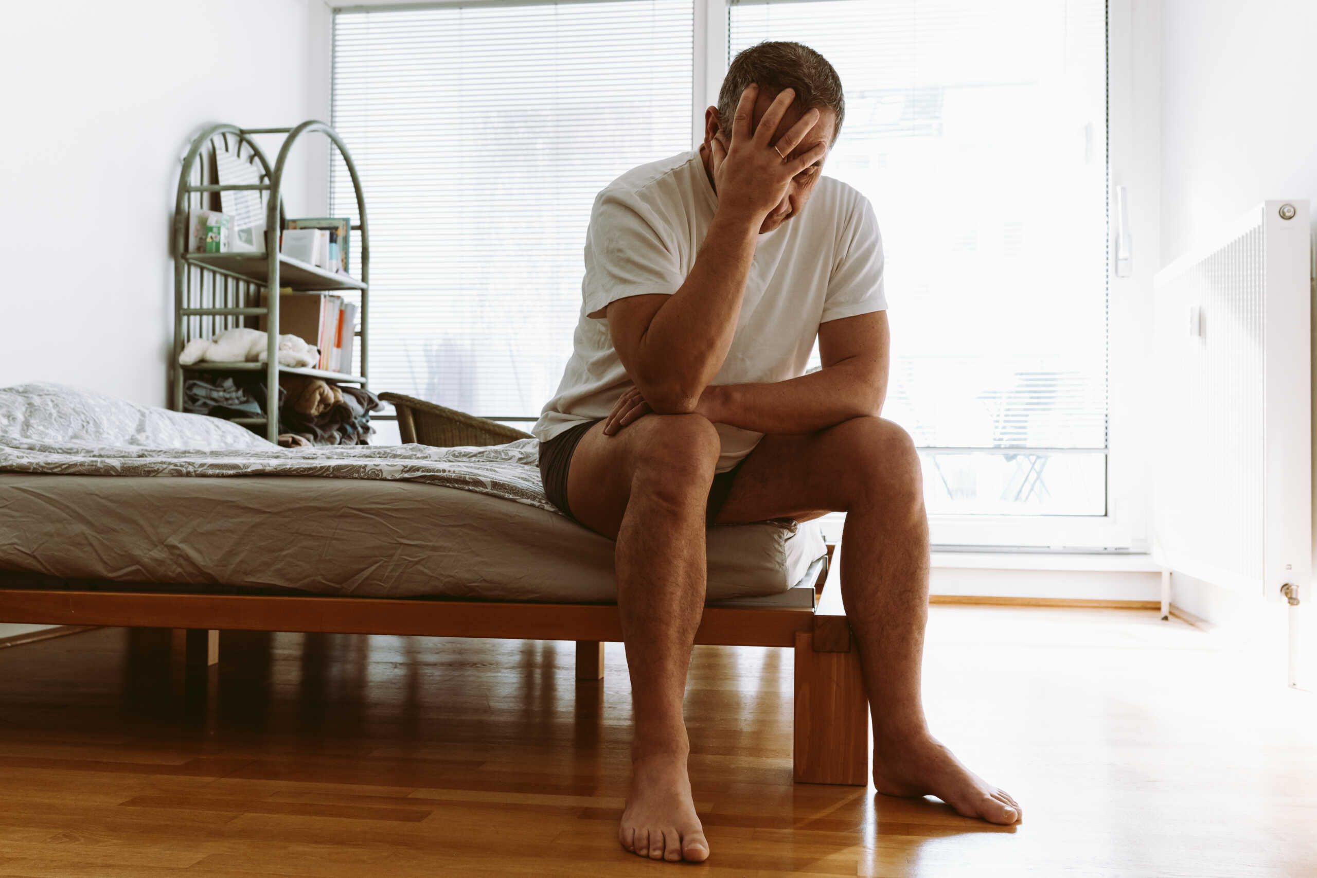 middle-aged man suffering from insomnia, depression, sits on bed with head down and covers face with hands.