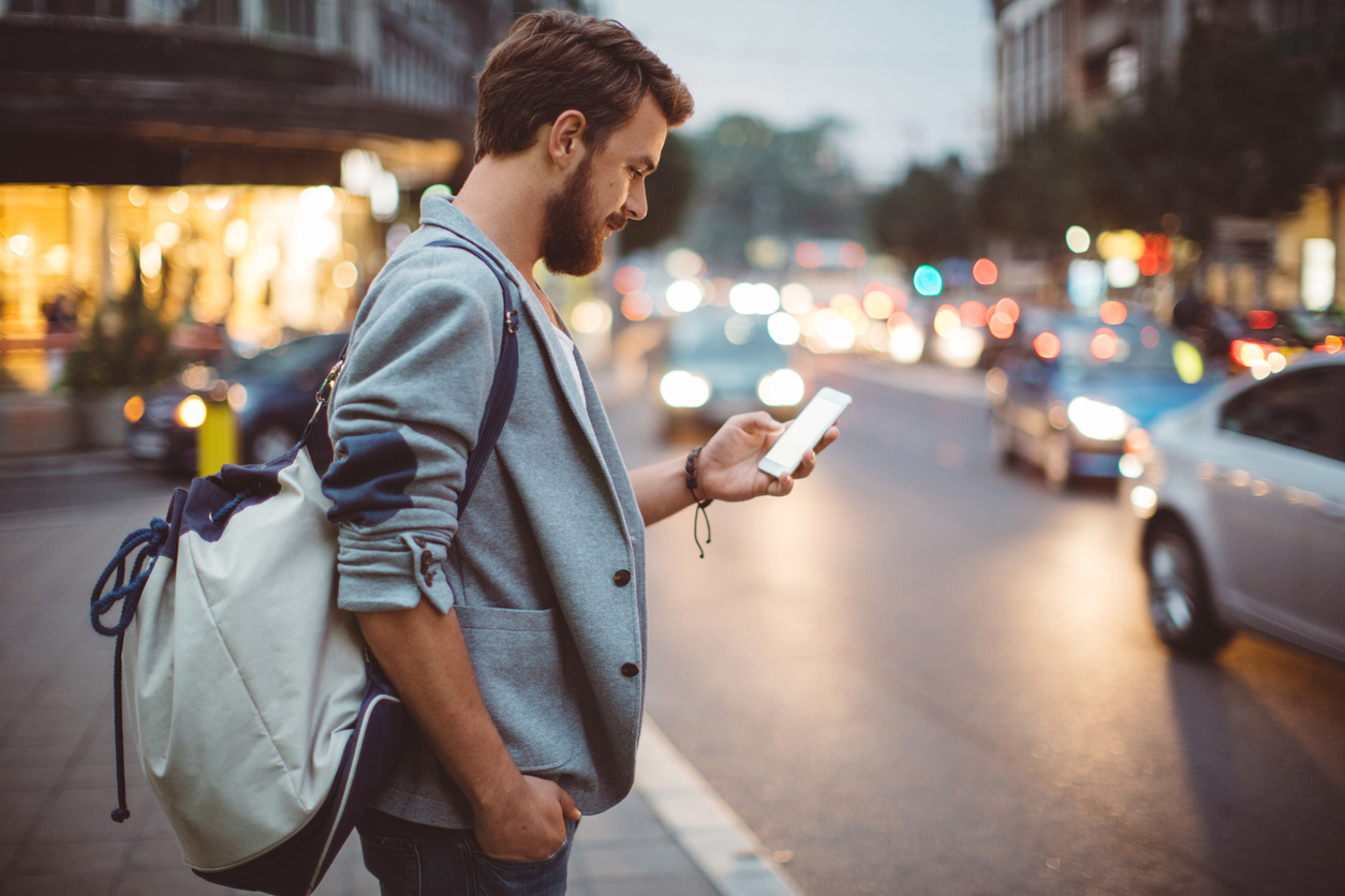 Young man on the street of big city , waiting taxi, chacking his smartphone, for news or new messages.  Or looking for map instructions.  Carry backpack on one shoulder.  Dusk time.  Casual dressed. Street lights in background.
