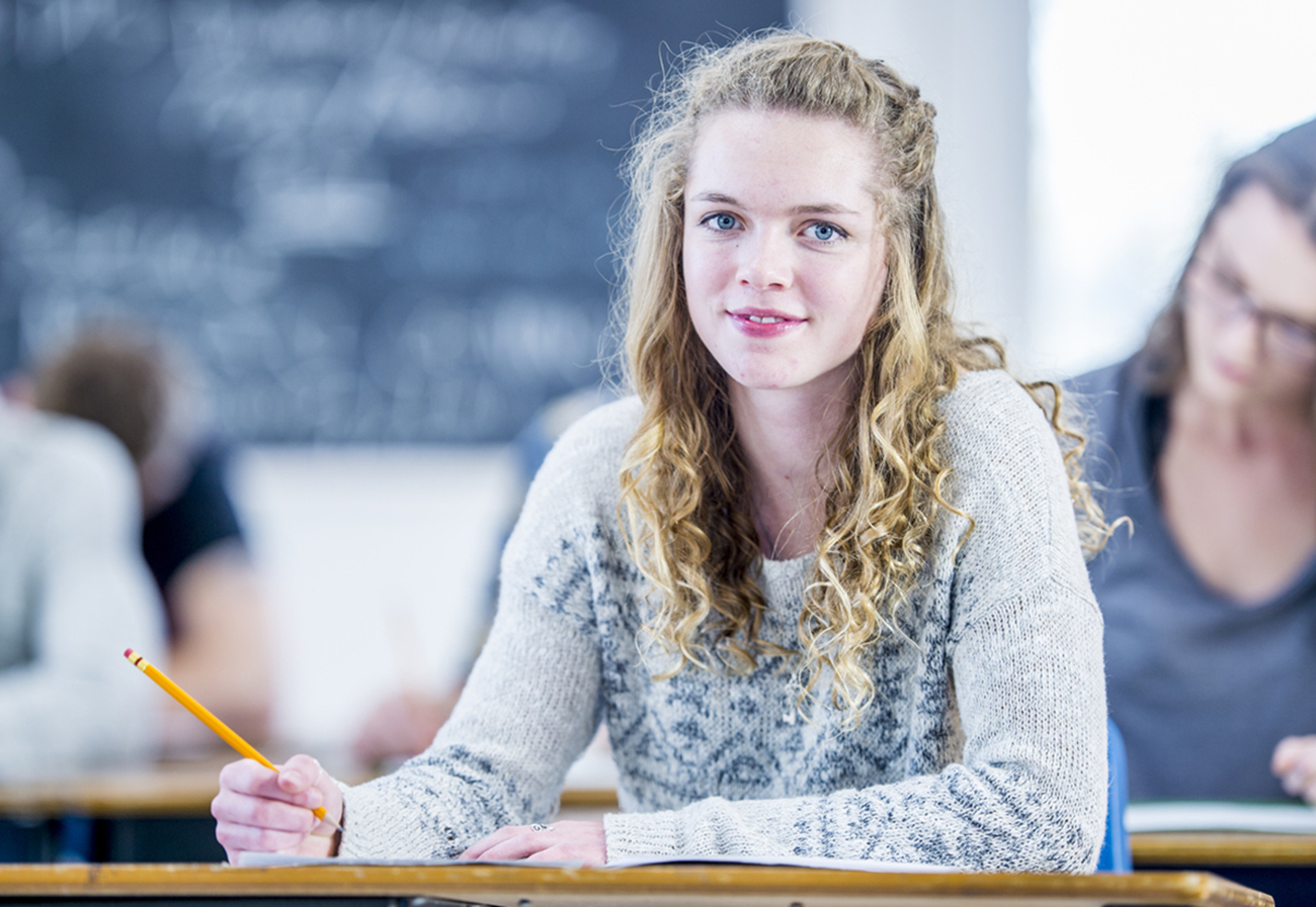 A multi-ethnic group of high school students are indoors in their classroom. They are all concentrating on writing a test. A Caucasian girl in the foreground is smiling at the camera.