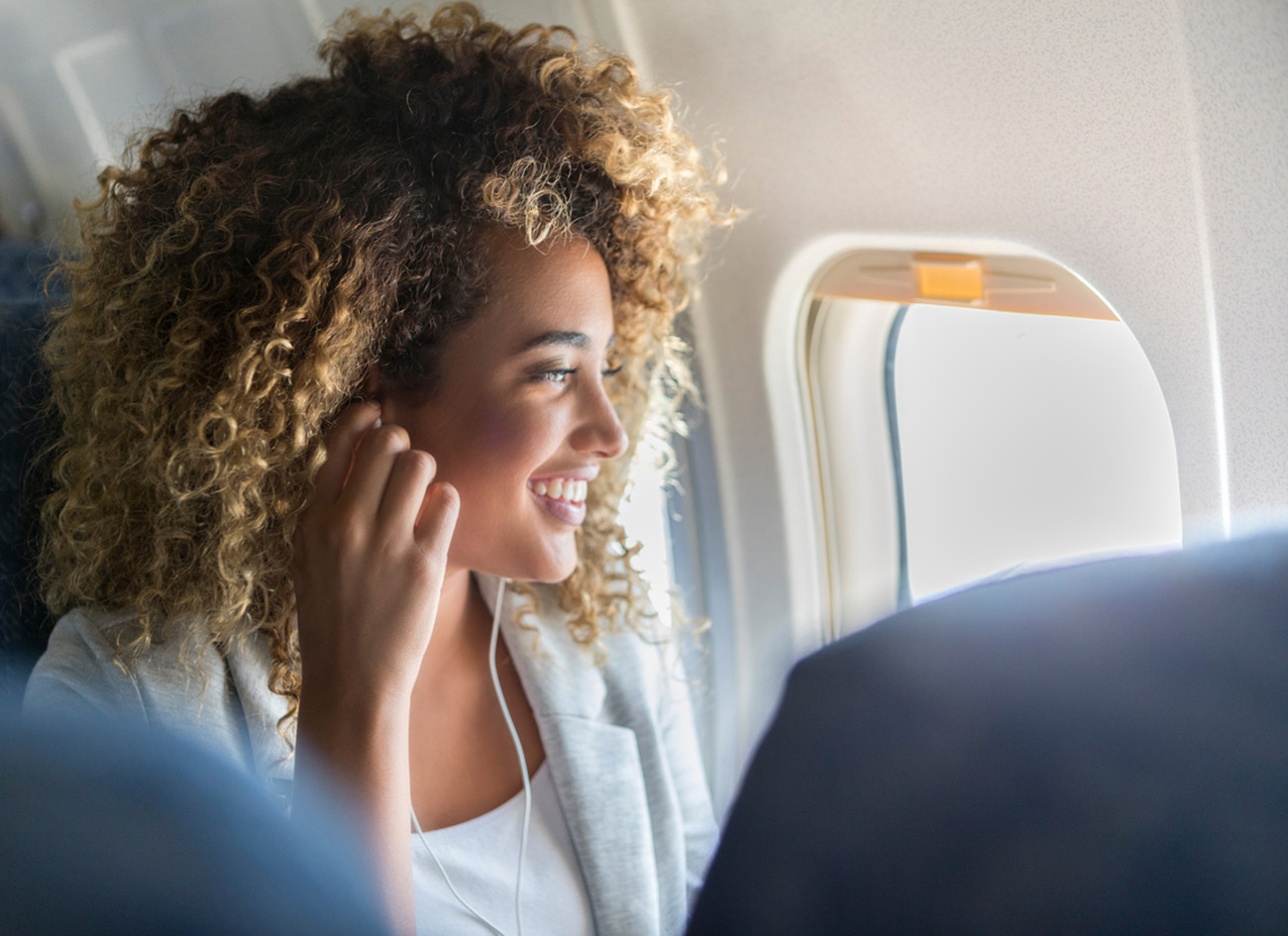 Attractive young woman wears earbuds to listen to music during a flight. She is sitting in a window seat gazing out of the window.