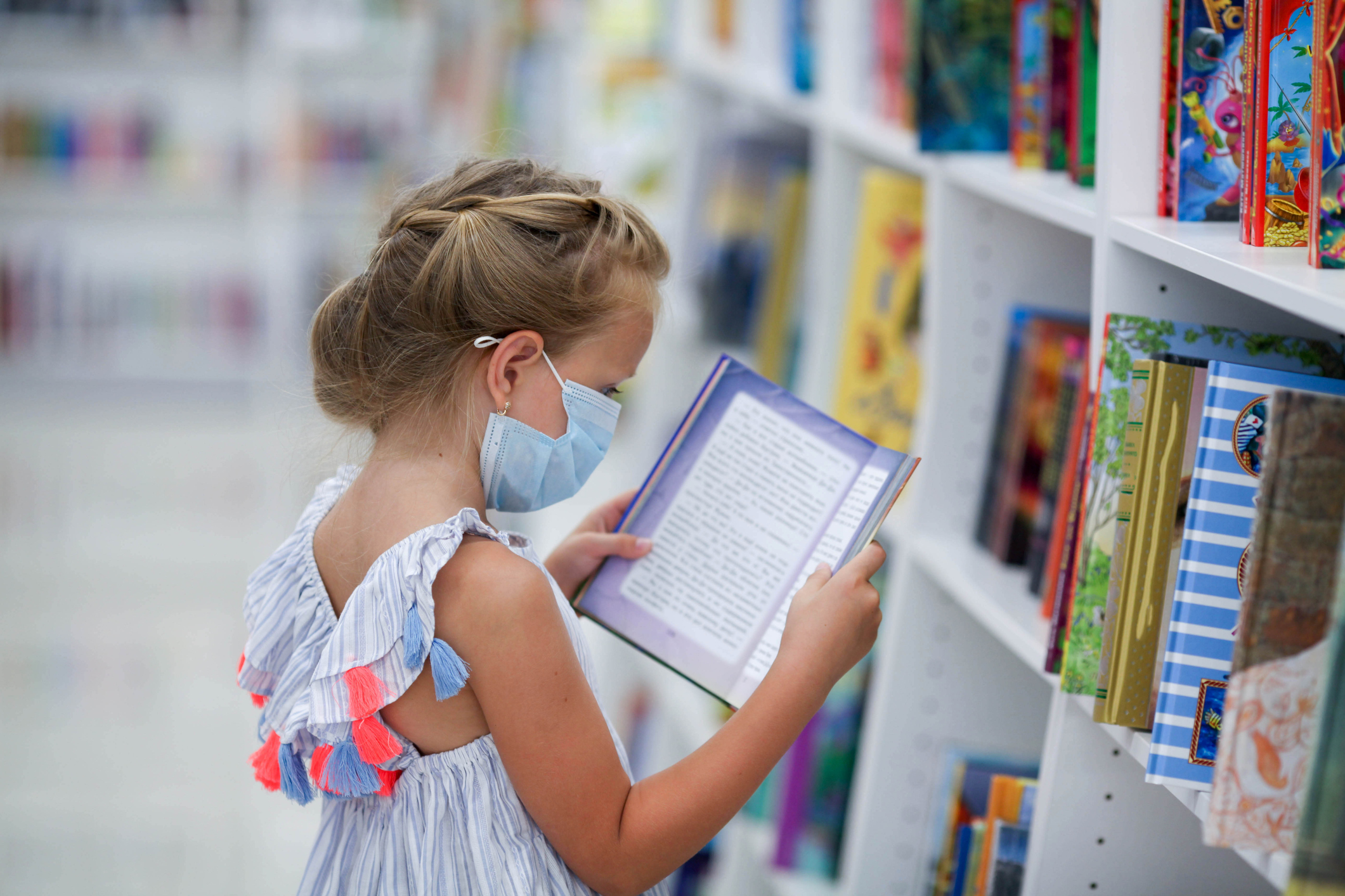 A little girl stands in a bookstore in a medical mask and reads. Child in the library.