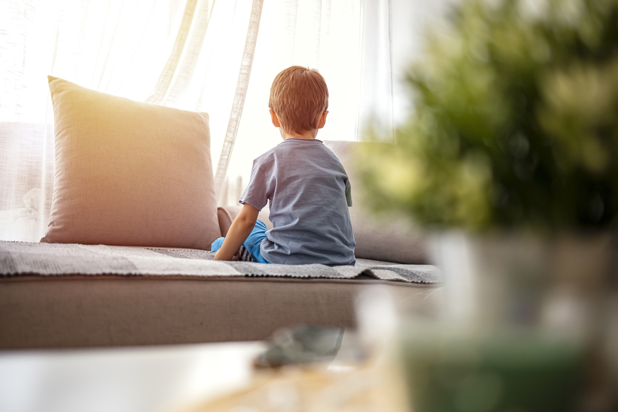 Boy from back, looking trough the window. Little boy sitting on sofa at home. Child autism. Depressed little boy sitting on the sofa. Unhappy child  Alone at home. Upset.