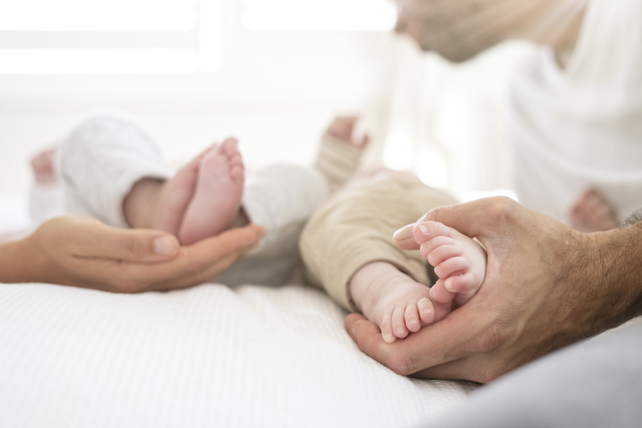 A new mother and father each hold the feet of their newborn fraternal twins laying on a bed. Shot is focused on feet and hands only.