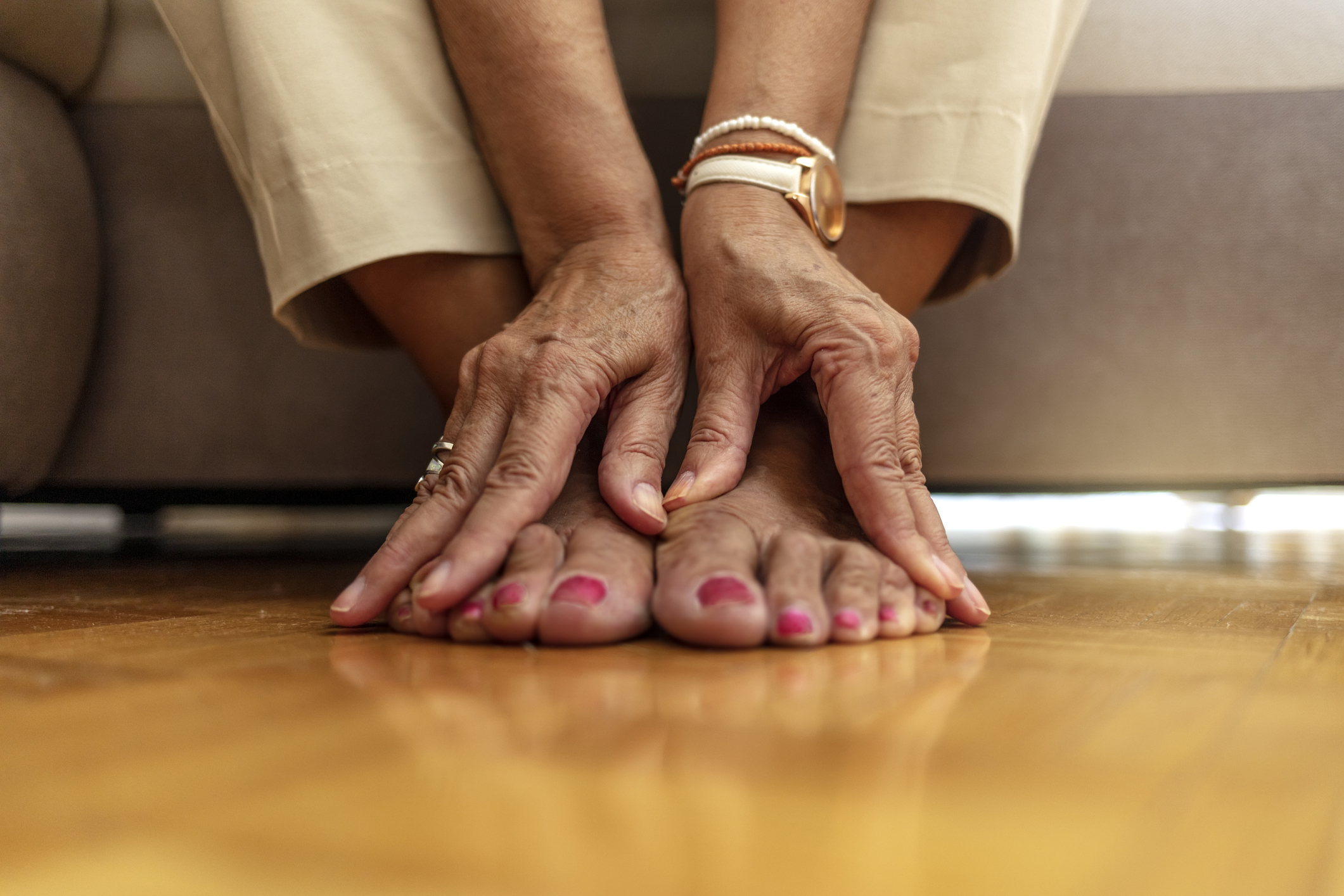 Close up of senior woman hands touching legs with varicose veins, sitting on sofa at home. Woman suffering from ankle pain. An older woman massages her feet to relieve the pain caused by arthritis.