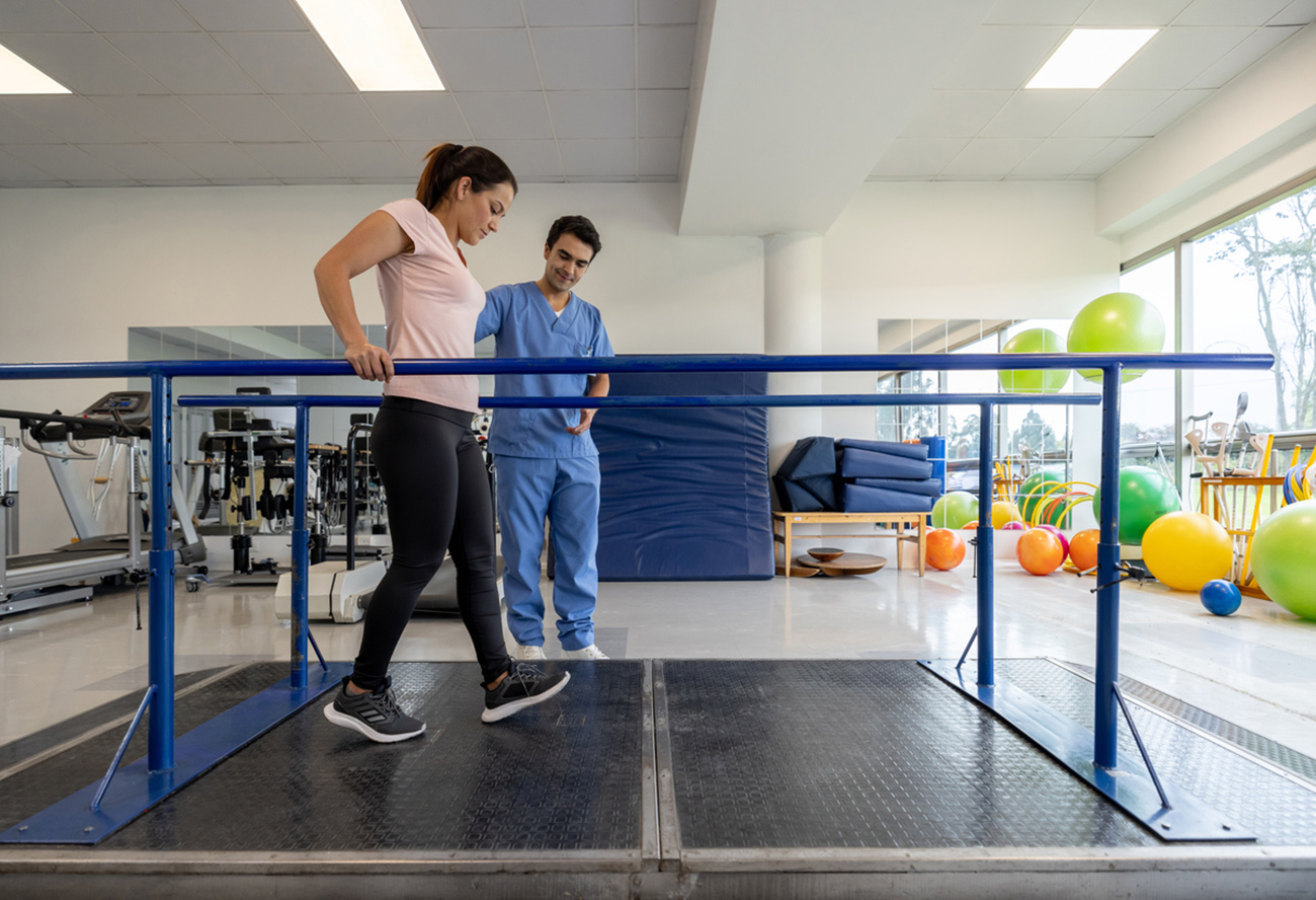 Latin American woman in physical therapy walking on the parallel bars with the assistance of his therapist