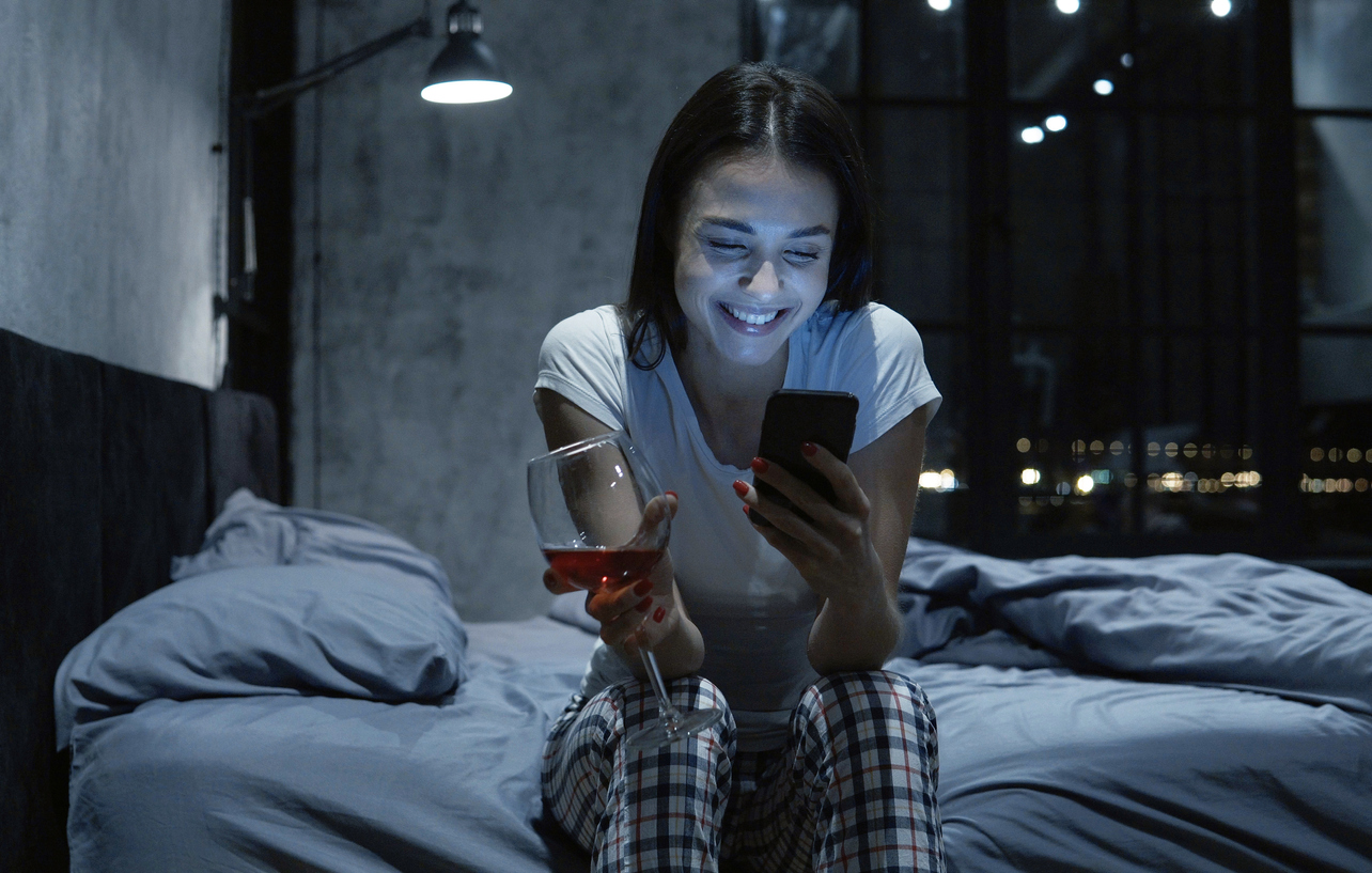 Social addicted beautiful young woman looking at smartphone, using online dating app, drinking wine, sitting on bed at night