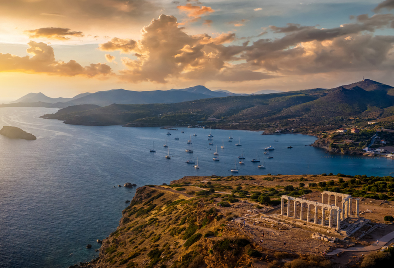 Panoramic view of the Temple of Poseidon at Cape Sounion at the edge of Attica, Greece, with moored sailboats in the bay during sunset time