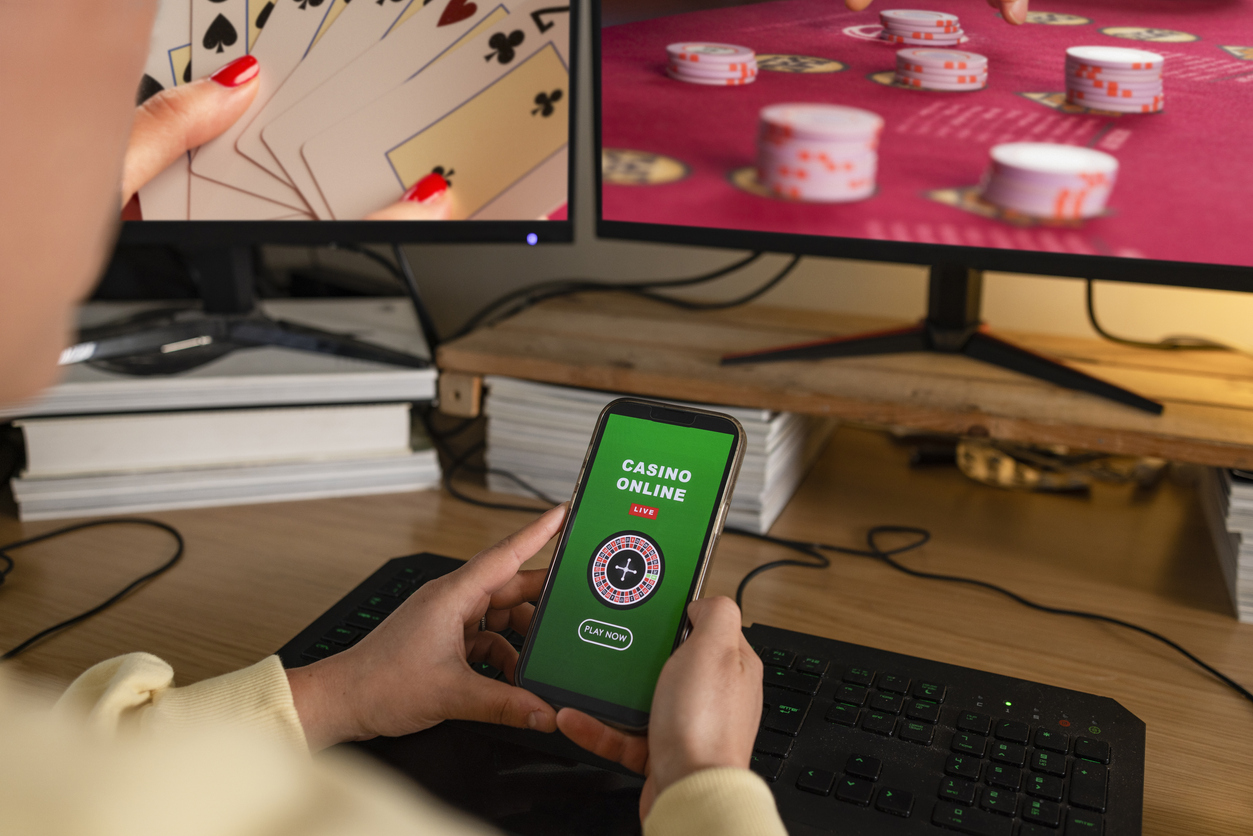 A rear-view shot of an unrecognisable young woman sitting at a desk with online gambling on her computer screens, she has a mouse and keyboard. She is also using her smart phone to gamble in an online casino.