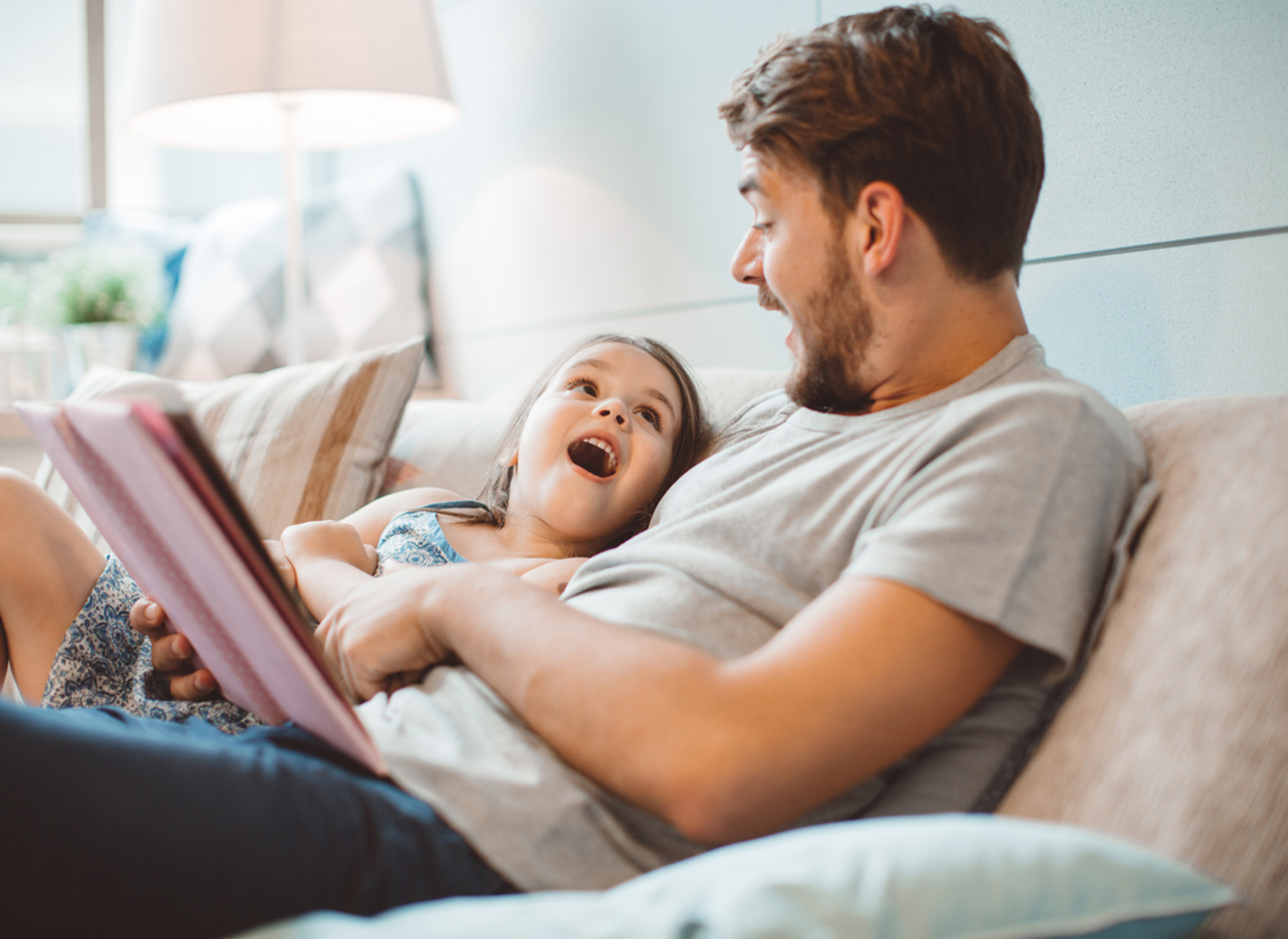 Father and daughter enjoying at home. Sitting on bed and reading book together.