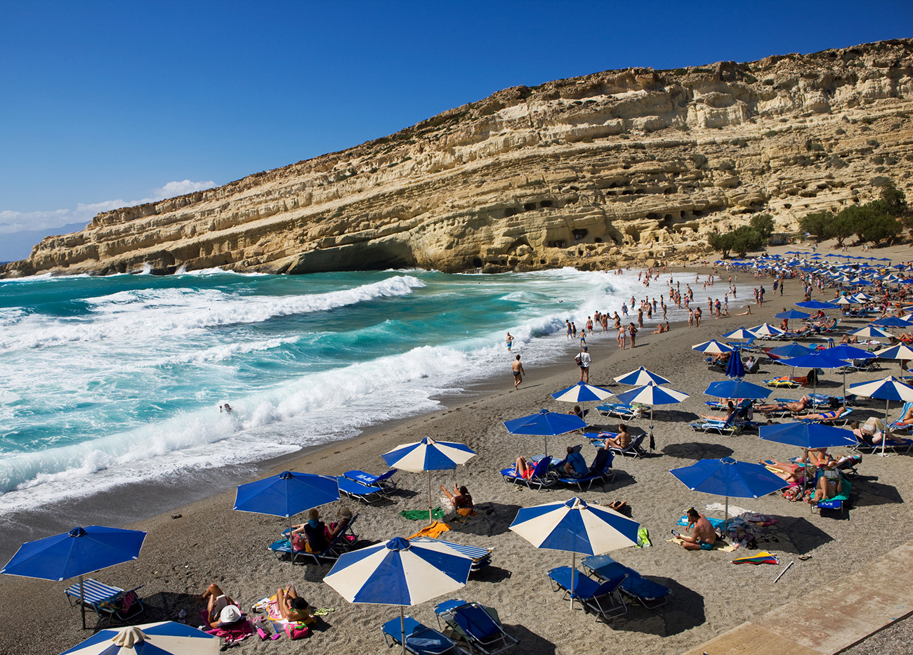 Matala, Greece - August 1, 2014: Many people relaxing at the famous beach of Matala on Crete. Matala is known as former place of pilgrimage for fans of the hippie era. There was a hippie community used to live in the caves above the beach and still there are some hippies around.