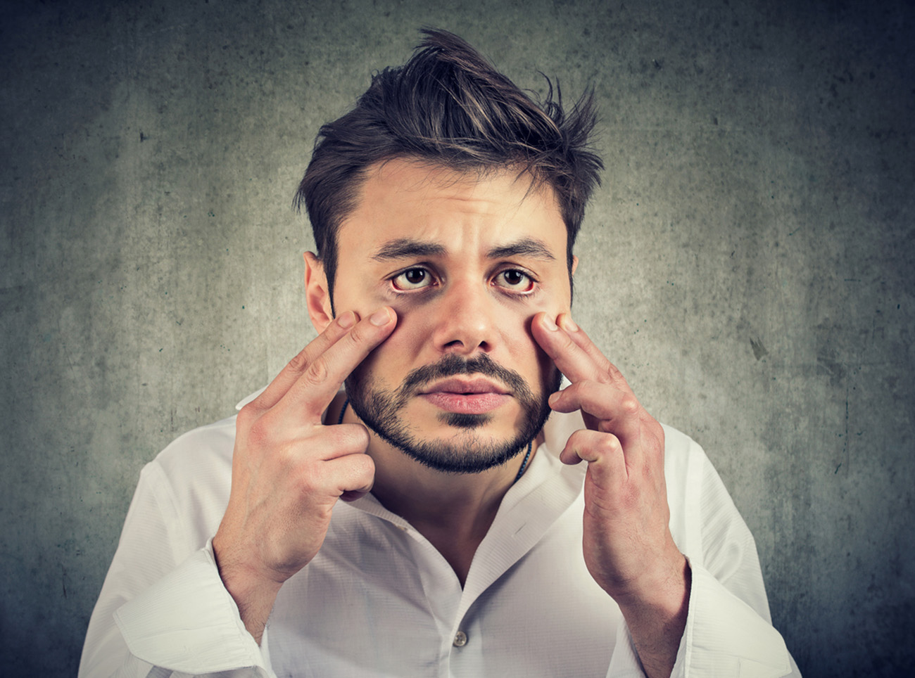 Young tired funny looking man making grimace and pullling down skin of eye bags being sick and unwell on gray background