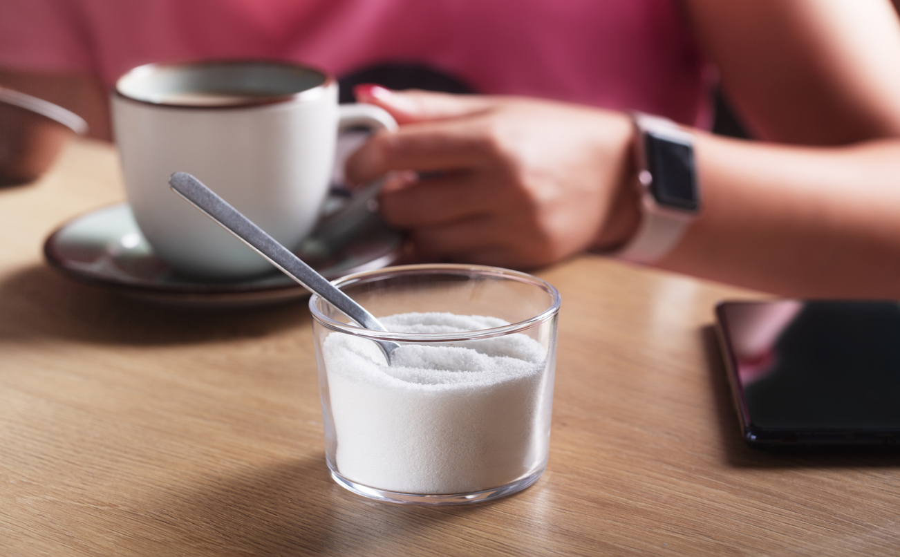 Woman with clock and phone holding a cup of coffee with milk and next to sugar substitute in a cafe