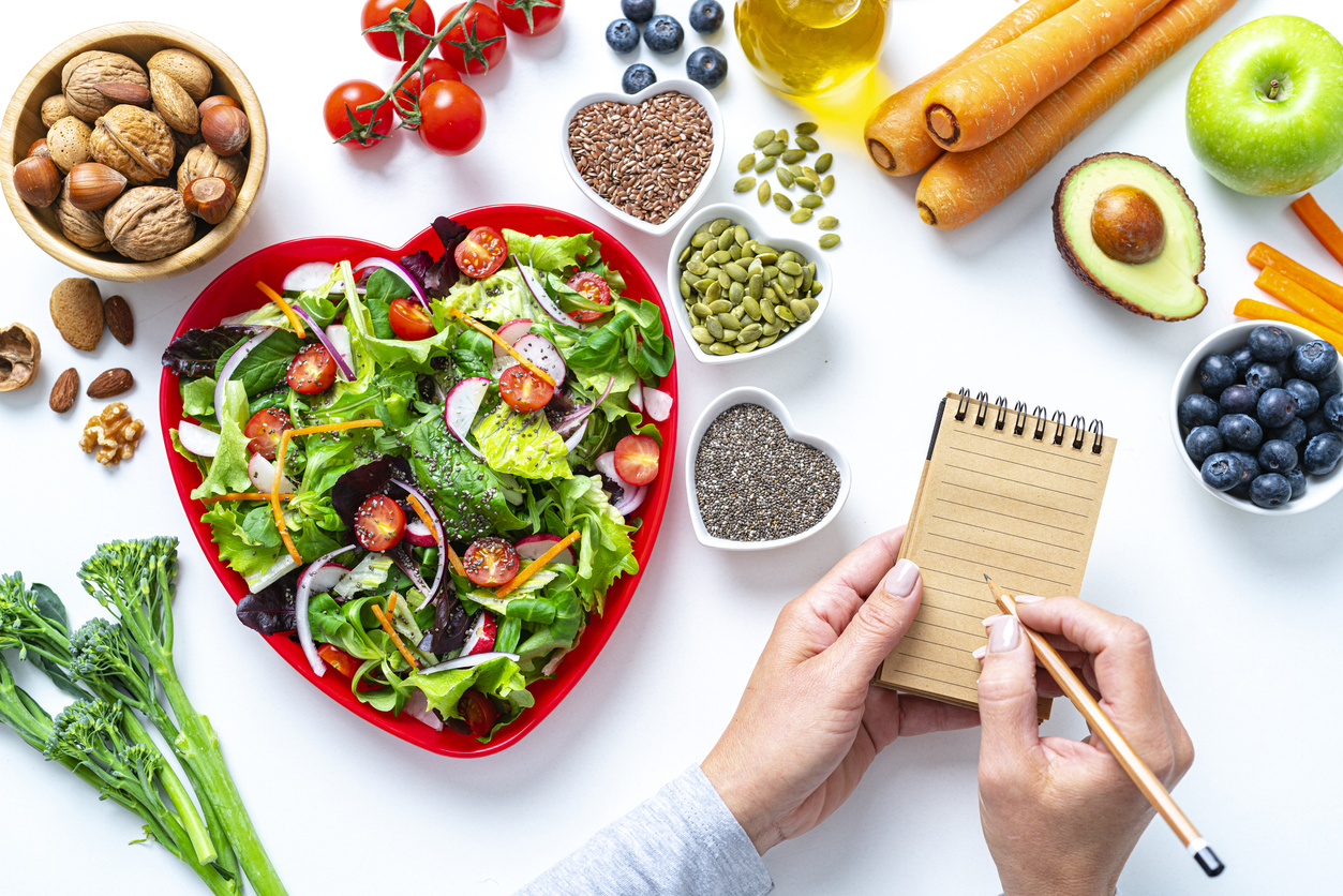 Overhead view of a red heart shape plate with healthy salad shot on white background. Water bottle, dumbbells and a tape measure are at the top right. Female hands are writing with a pencil on a note pad. Multi colored fresh fruits, vegetables, seeds and nuts are around the salad plate. High resolution 42Mp studio digital capture taken with SONY A7rII and Zeiss Batis 40mm F2.0 CF lens