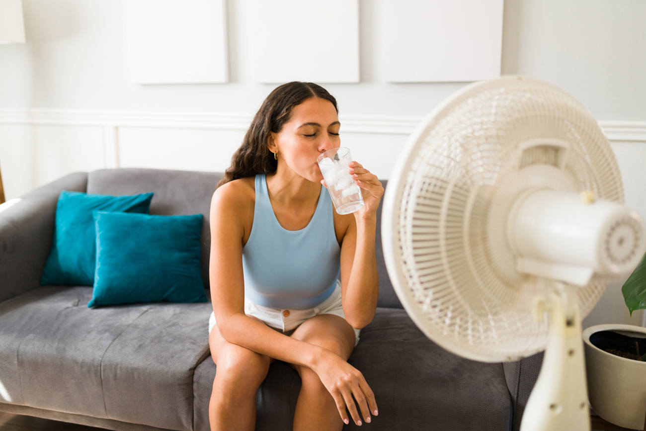 Happy woman trying to cool down drinking an ice cold drink and turning on the electric fan at home