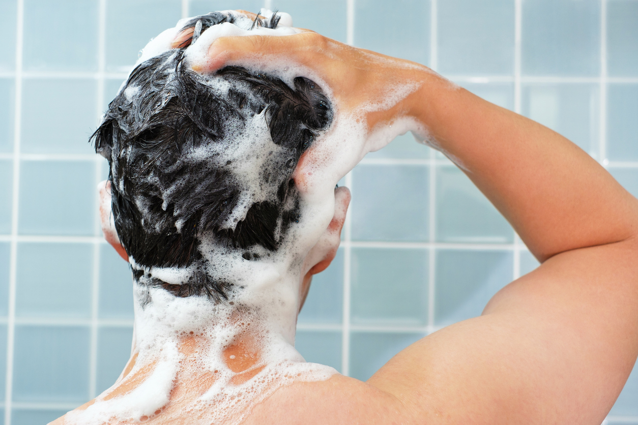 Male hands wash their hair with shampoo and foam on a blue background, rear view.