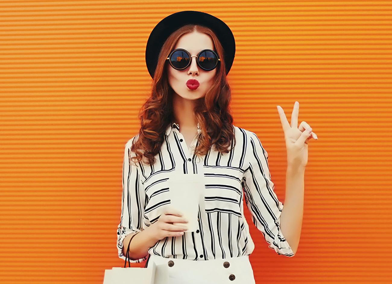 Portrait of beautiful young woman model with shopping bags and cup of coffee blowing her lips sending air kiss wearing white striped shirt, black round hat on orange background