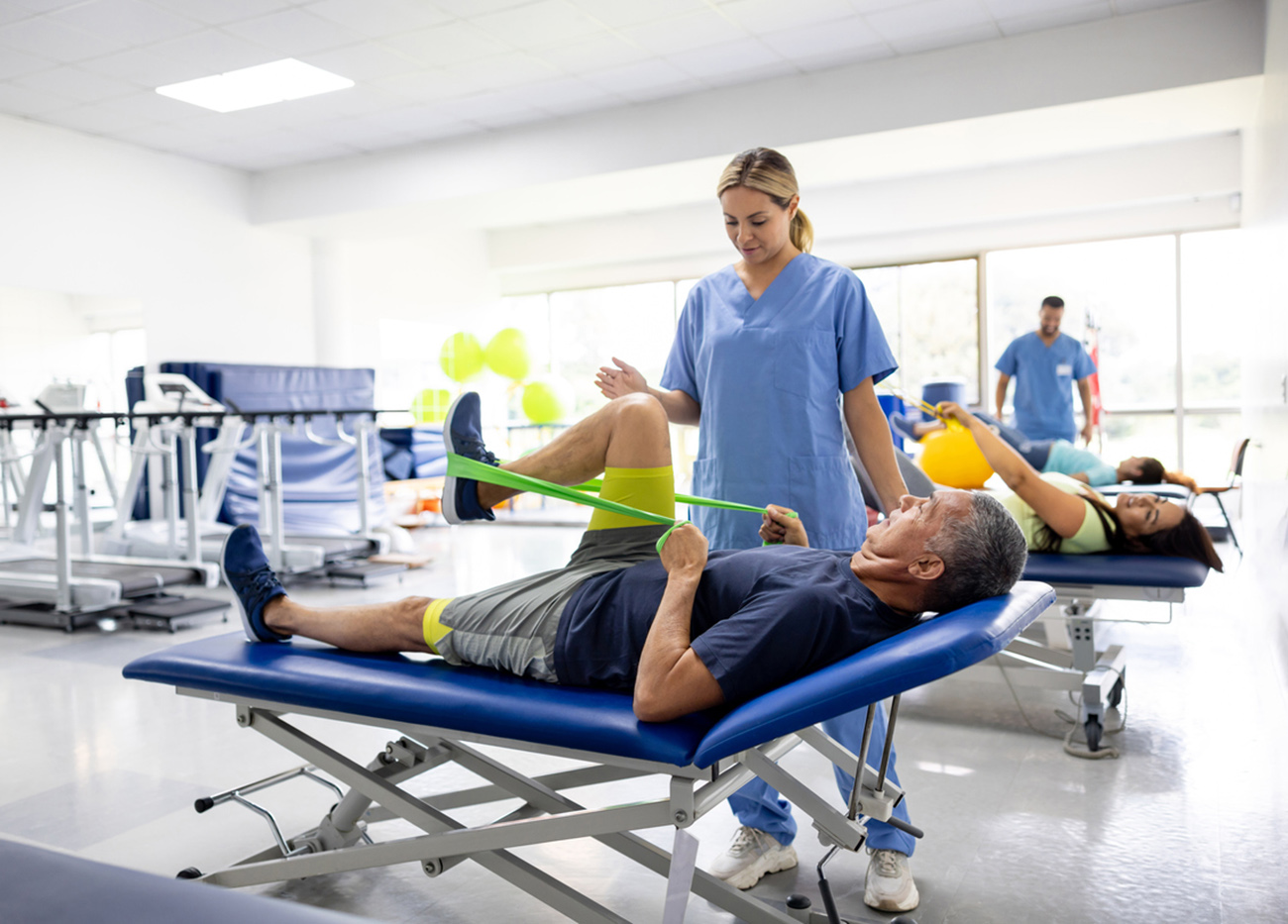 Mature Latin American man doing physical therapy exercises using a stretch band with the assistance of his therapist