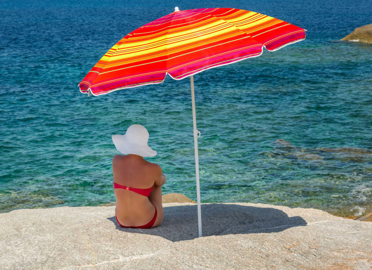 Woman in red bikini and large white floppy hat sitting under a brightly coloured red and yellow striped parasol on a rock and looking out across the blue Mediterranean sea