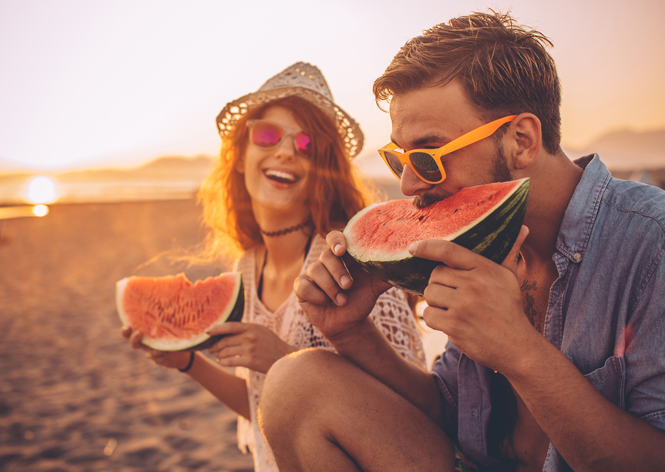 Hipster couple sitting on the beach and eating watermelon. Wearing casual summer clothing, hat and sunglasses. Enjoying in sunset by the sea.