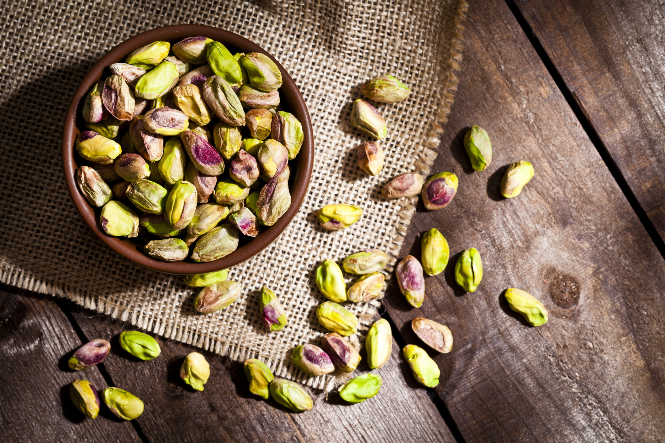 Top view of a brown bowl filled with organic pistachios shot on rustic wood table. Some almonds are out of the bowl on a burlap. Predominant colors are green and brown. DSRL studio photo taken with Canon EOS 5D Mk II and Canon EF 100mm f