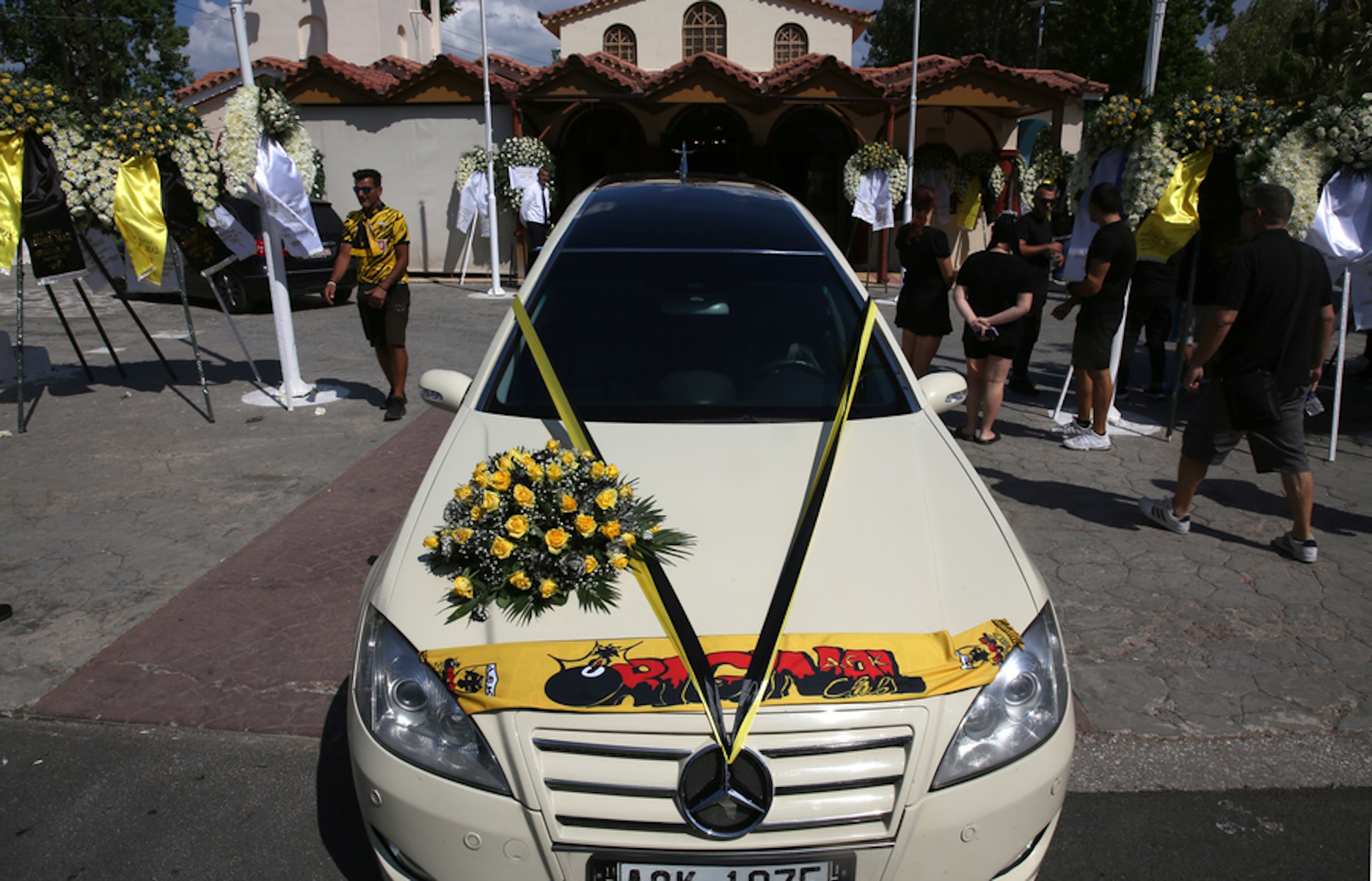 Νεκροφόρα με κίτρινα λουλούδια και το κασκόλ της ΑΕΚ βρίσκεται έξω από τον Ιερό Ναό του  Αγίου Κωνσταντίνου για την κηδεία του αδικοχαμένου Μιχάλη Κατσουρή, τον  φίλαθλο της ΑΕΚ που βρήκε το θάνατο από τη δολοφονική επίθεση της περασμένης Δευτέρας, παραμονή του ευρωπαϊκού αγώνα με τη Ντιναμό Ζάγκρεμπ έξω από το γήπεδο της Νέας Φιλαδέλφειας, Ελευσίνα, Παρασκευή 11 Αυγούστου 2023. Η εξόδιος ακολουθία θα πραγματοποιηθεί στις 5 το απόγευμα και η οικογένεια του Μιχάλη ζήτησε –αντί στεφάνων- να κατατεθούν δωρεές για το ίδρυμα «Ελπίδα». Το μήνυμα της οικογένειας: «Ο Μιχάλης μας ήταν παιδί ευαίσθητο, καλόκαρδο που βοήθησε με αυτοθυσία στις φωτιες, βοηθούσε άστεγους και είχε μεγάλη πίστη στο Θεό. Γι' αυτό η οικογένεια του, αποφασίσαμε αντί στεφάνων να γίνουν δωρεές για μικρά παιδιά, υπέρ του Σωματείου "ΕΛΠΙΔΑ -ΣΥΛΛΟΓΟΣ ΦΙΛΩΝ ΠΑΙΔΙΩΝ ΜΕ ΚΑΡΚΙΝΟ" στη μνήμη του αδικοχαμένου μας». ΑΠΕ-ΜΠΕ