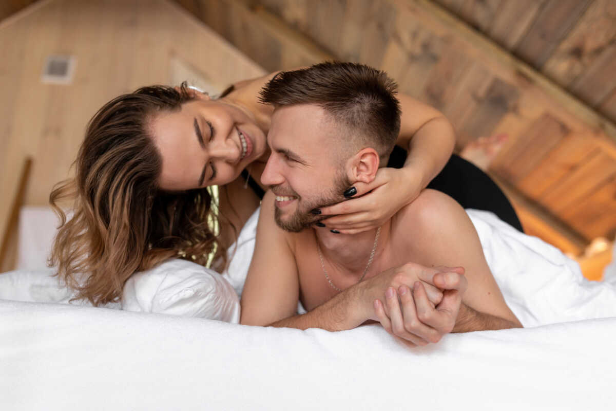 Ð¡harming long-haired brown-haired woman playfully embraces her beloved, they are lying in bed. Safe sex. Beauty and youth. Lifestyle. Romantic date. Softness. Free relationship. Family life.