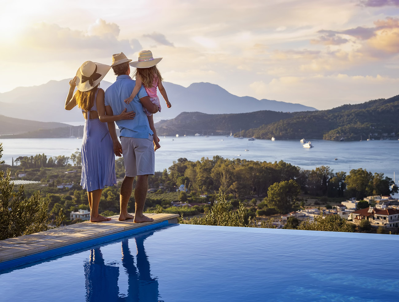 A family on summer holidays stands by the swimming pool and enjoys the beautiful sunset behind the mountains and the sea
