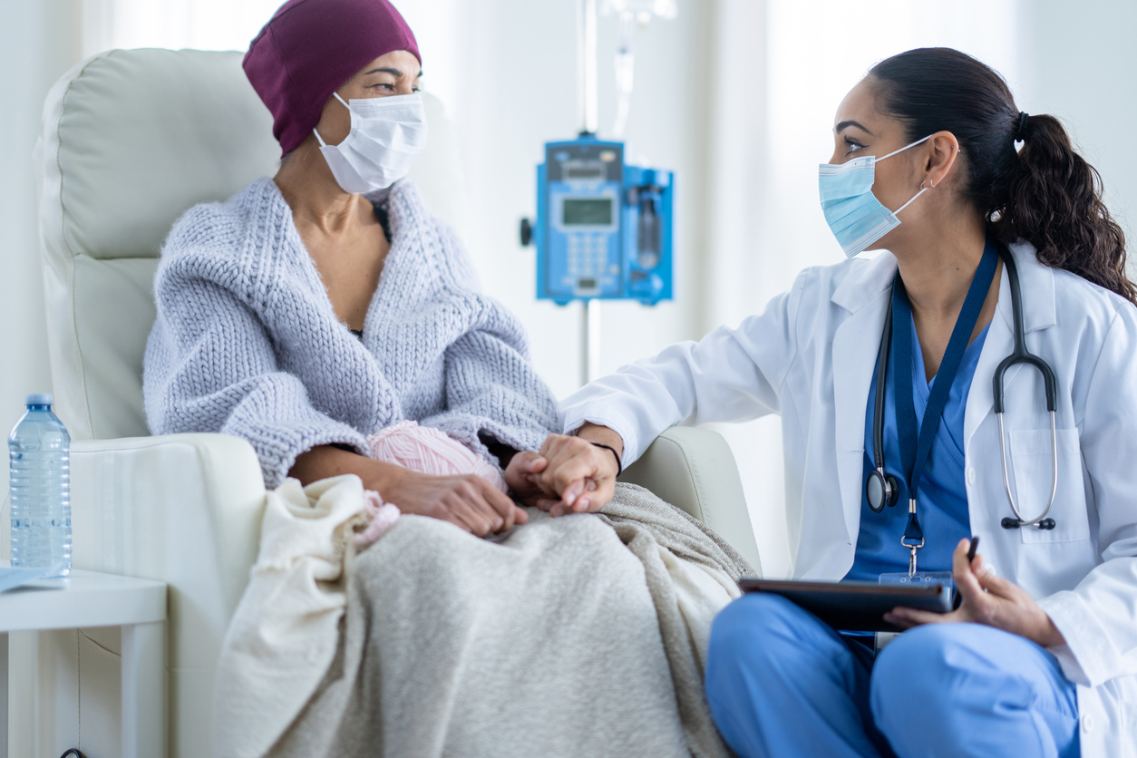 A senior woman battling cancer sits in a high back chair as she talks with her Oncologist during a Chemotherapy treatment.  The patent is dressed casually in a sweater and has a blanket and headscarf on to keep her warm.