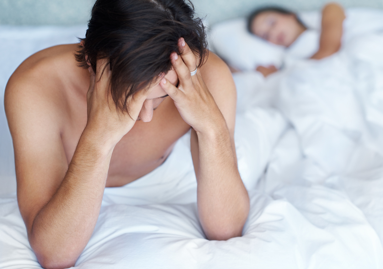 A young man sitting on the edge of his bed while his wife sleeps in the background