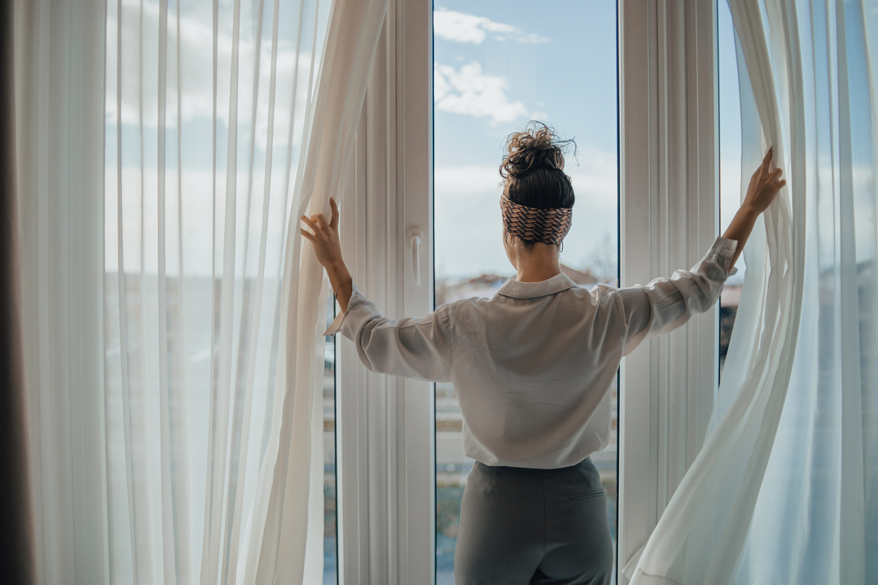 One young woman opens the curtains in the room in the morning at sunrise, she is dressed in a white shirt, ready for work and daily duties, morning routine