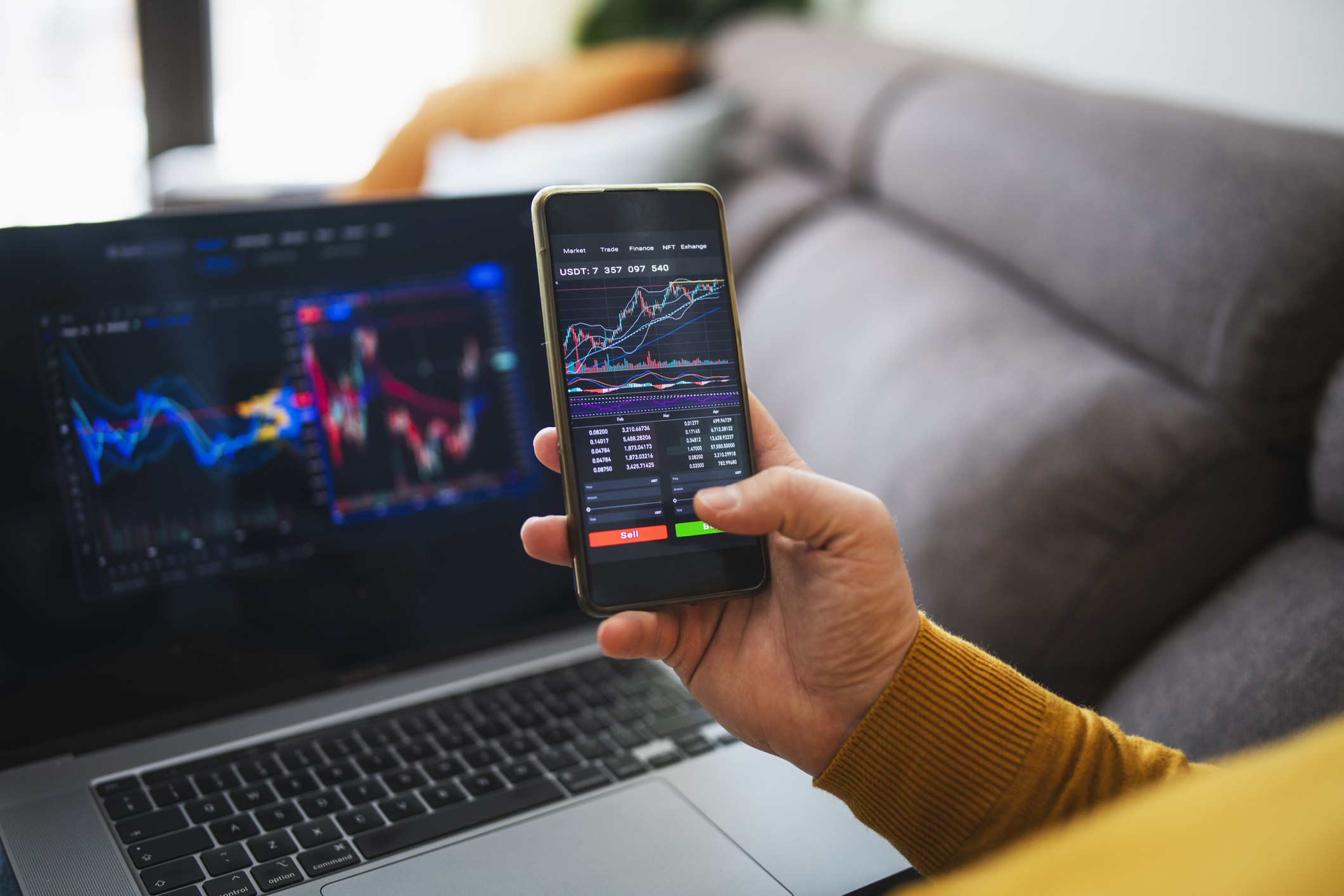 Rear view of man at home using laptop and phone to trade with cryptocurrency.