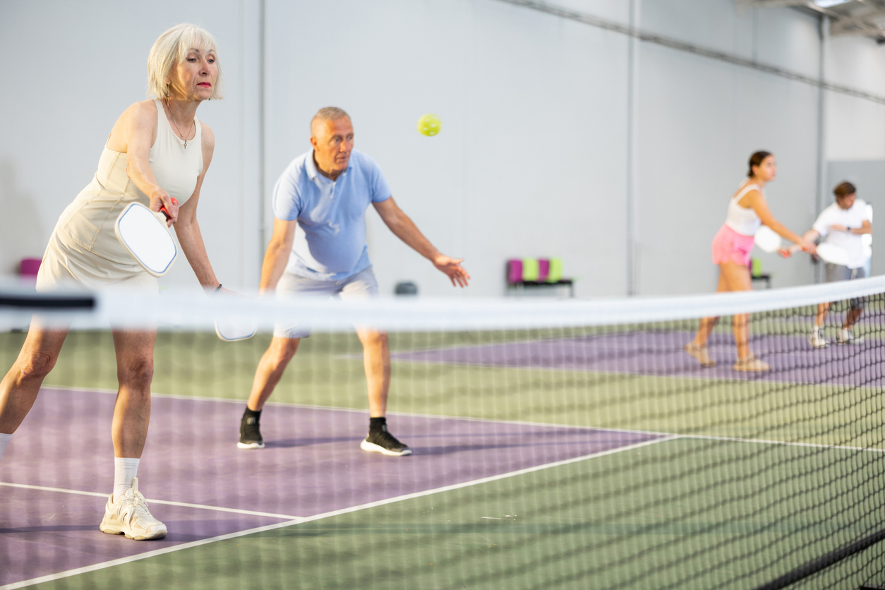 Focused positive elderly woman playing friendly pickleball match in team with male partner on indoors court. Concept of concentration in competition